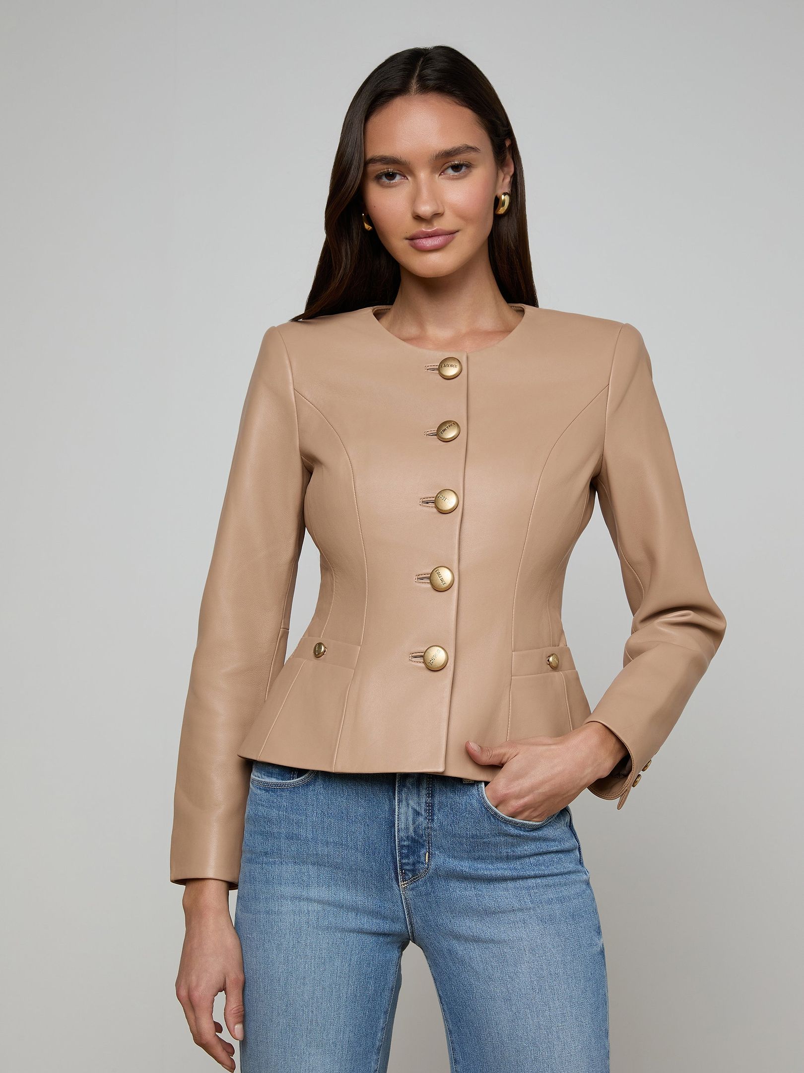 A woman wearing the Mariella Leather Jacket—a fitted, camel-colored chic leather piece with gold buttons—paired with blue jeans, stands against a plain, light background with a neutral expression.