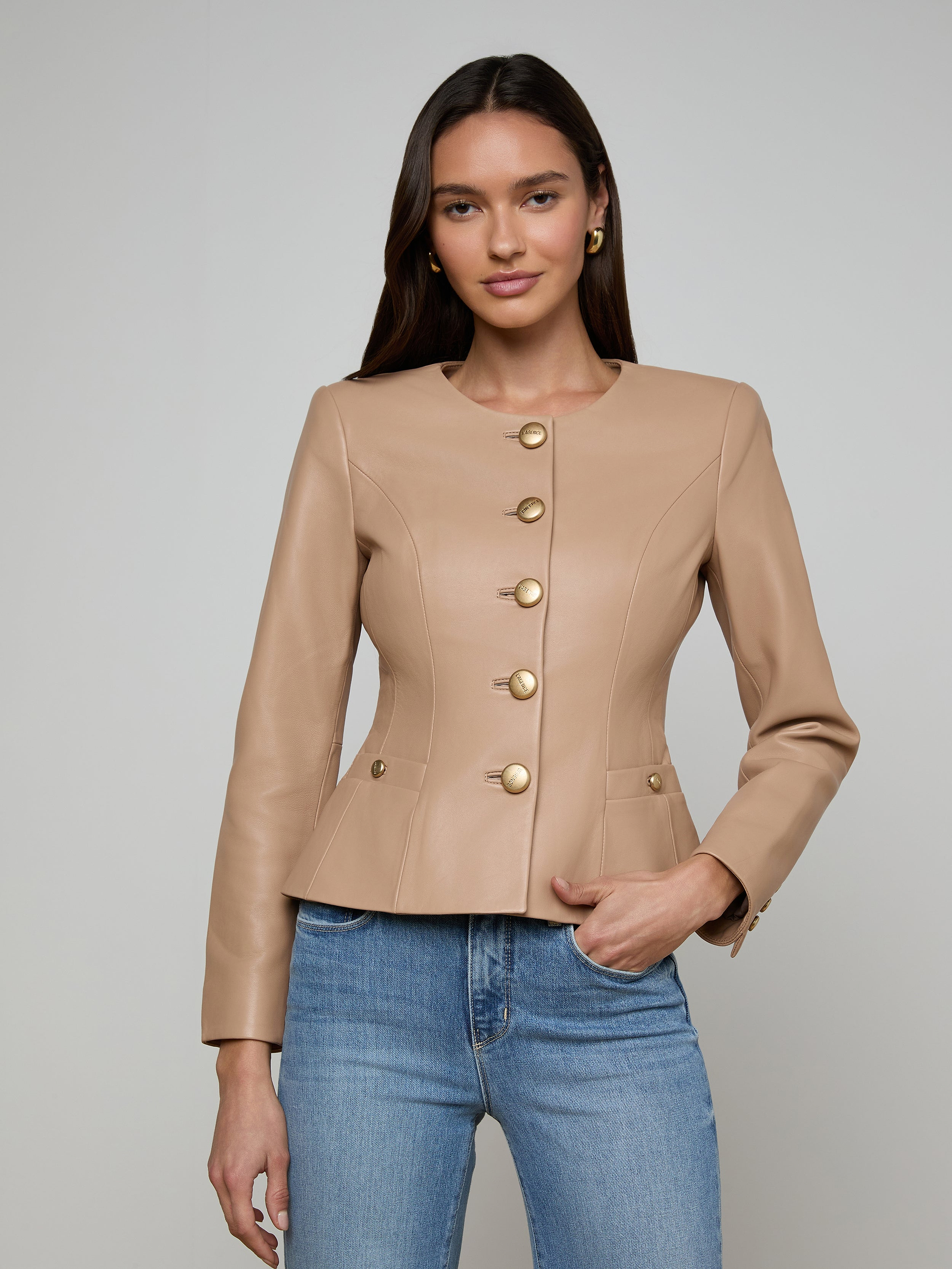 A woman wearing the Mariella Leather Jacket—a fitted, camel-colored chic leather piece with gold buttons—paired with blue jeans, stands against a plain, light background with a neutral expression.