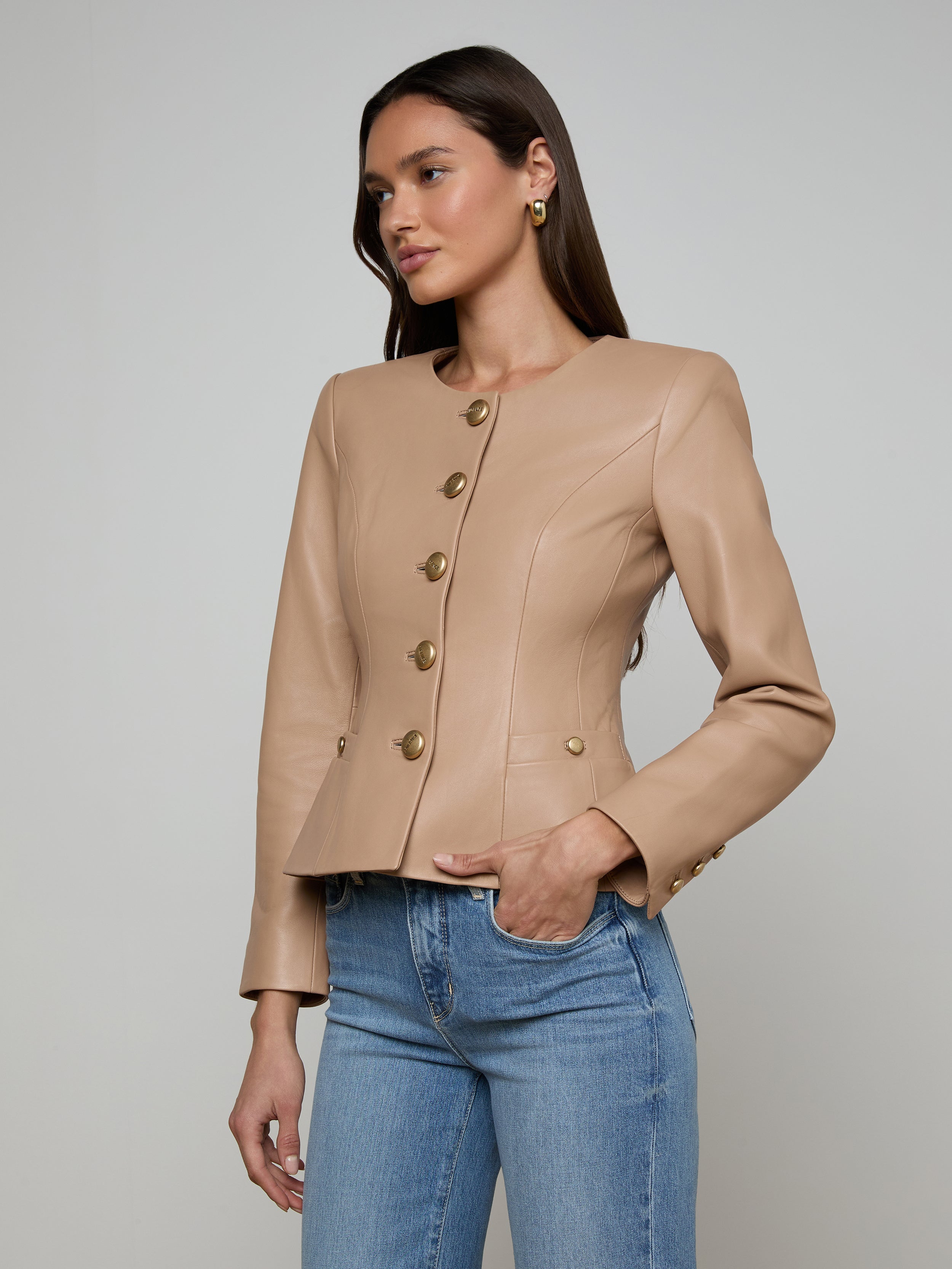 A woman models the Mariella Leather Jacket in a chic collarless design and cappuccino shade, styled with blue jeans. She stands against a plain light background, gazing slightly to the side with one hand in her pocket.