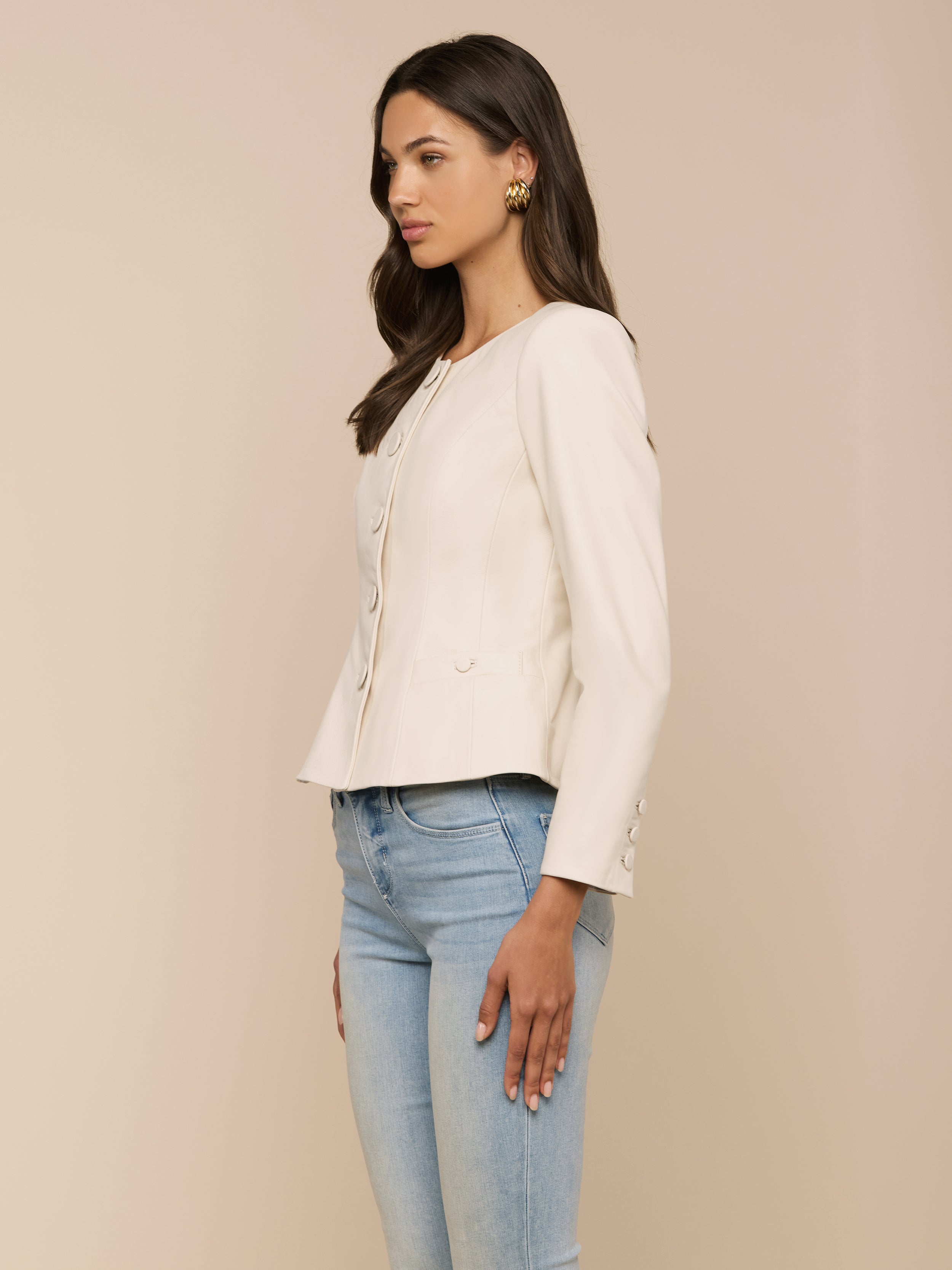 A woman with long brown hair stands sideways against a plain light background, styled in a cream-colored, button-up long-sleeve blouse and light blue jeans. She accessorizes with gold earrings and completes her outfit with the Mariella Leather Jacket for a chic finish.