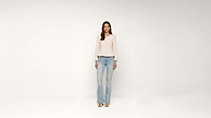Woman stands against a plain white background, wearing a light-colored blouse, blue jeans, and white open-toe shoes. She has long brown hair and her arms rest naturally by her sides.