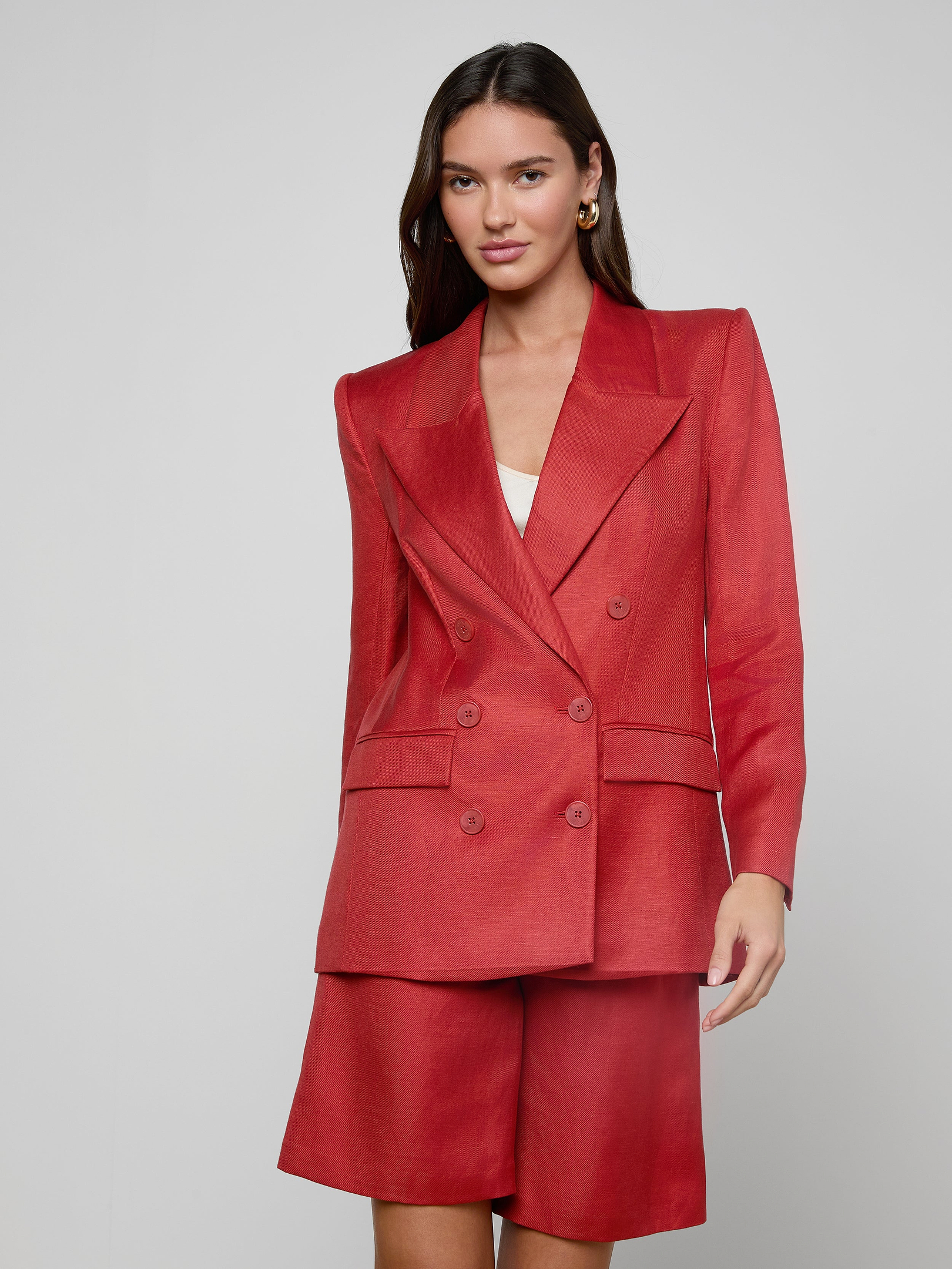 A woman wears the Cambrie Oversized Blazer in red linen, paired with matching knee-length shorts, standing against a plain light background. She has long brown hair and a slight smile.