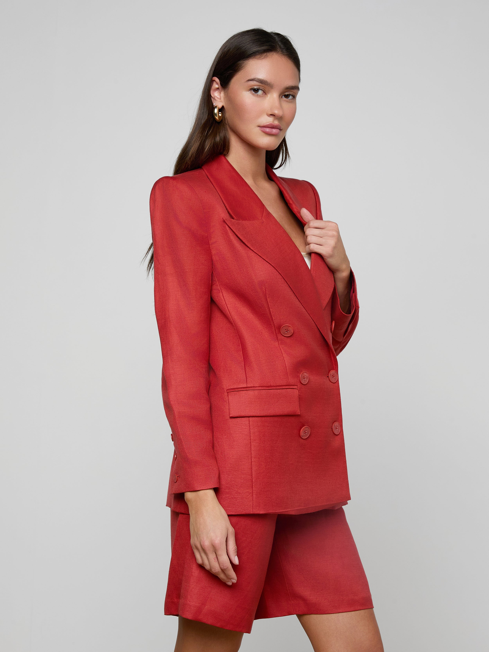 A woman with long brown hair and gold earrings stands against a light background, wearing the Cambrie Oversized Blazer in red paired with matching tailored shorts, her expression neutral.