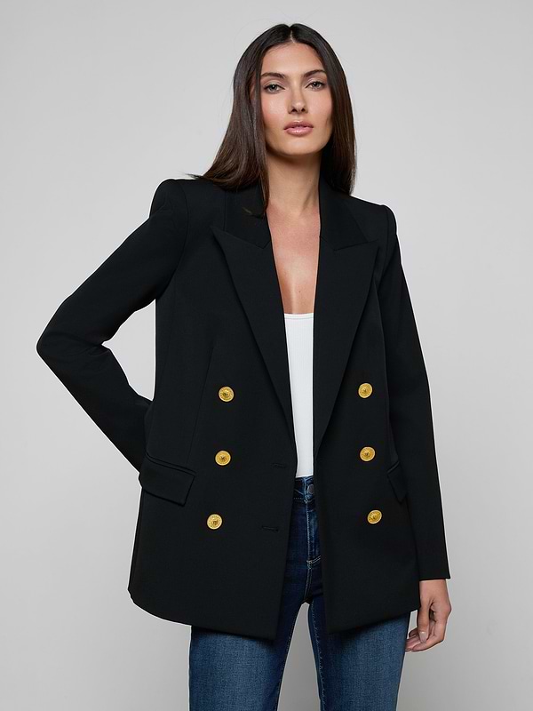 A woman wears the Cambrie Oversized Blazer, crafted from stretch suit fabric in black with gold buttons, layered over a white top and blue jeans as she stands against a plain light background.