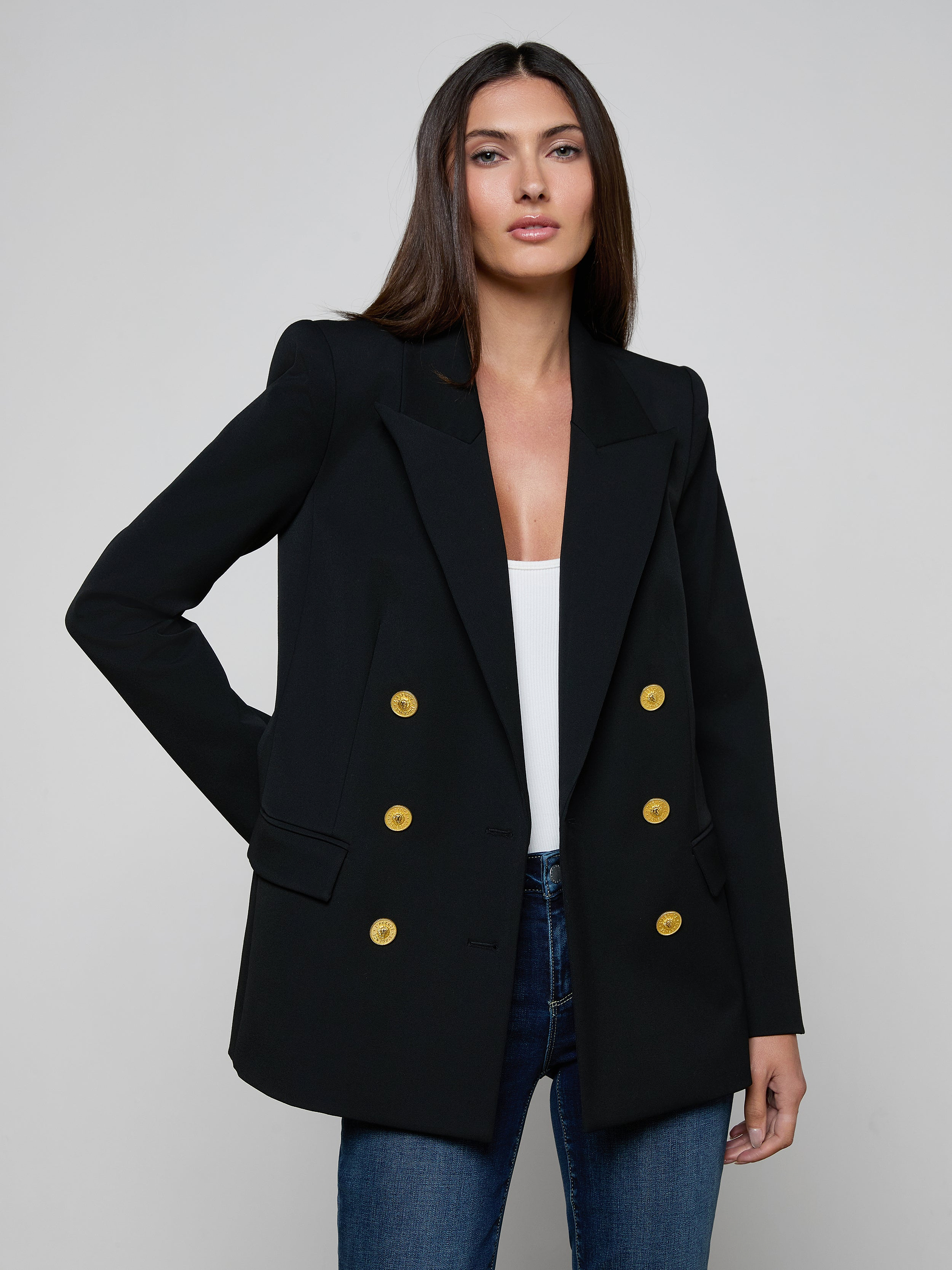 A woman wears the Cambrie Oversized Blazer, crafted from stretch suit fabric in black with gold buttons, layered over a white top and blue jeans as she stands against a plain light background.