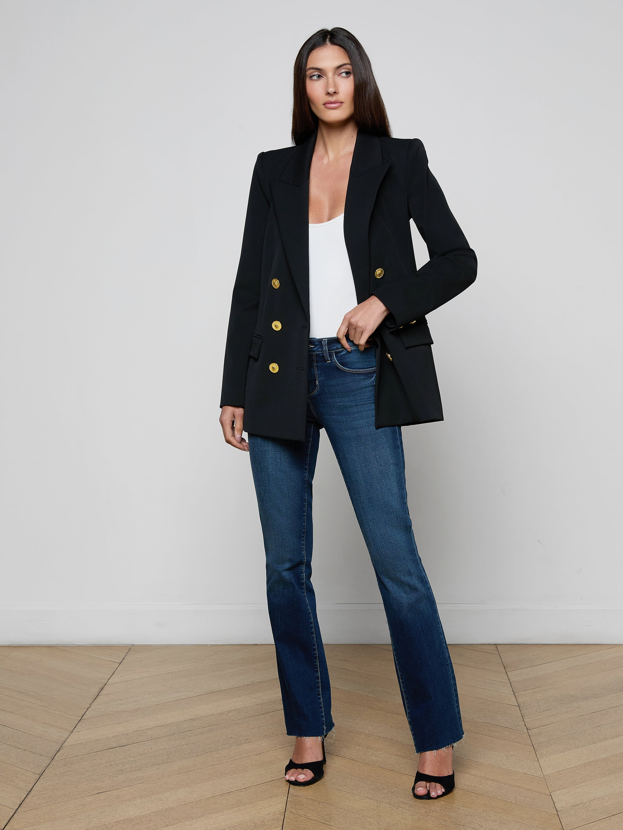A woman stands facing forward on a plain white background, wearing the Cambrie Oversized Blazer—an oversized black blazer with gold buttons crafted from stretch suit fabric—paired with a white top, blue jeans, and black heels.