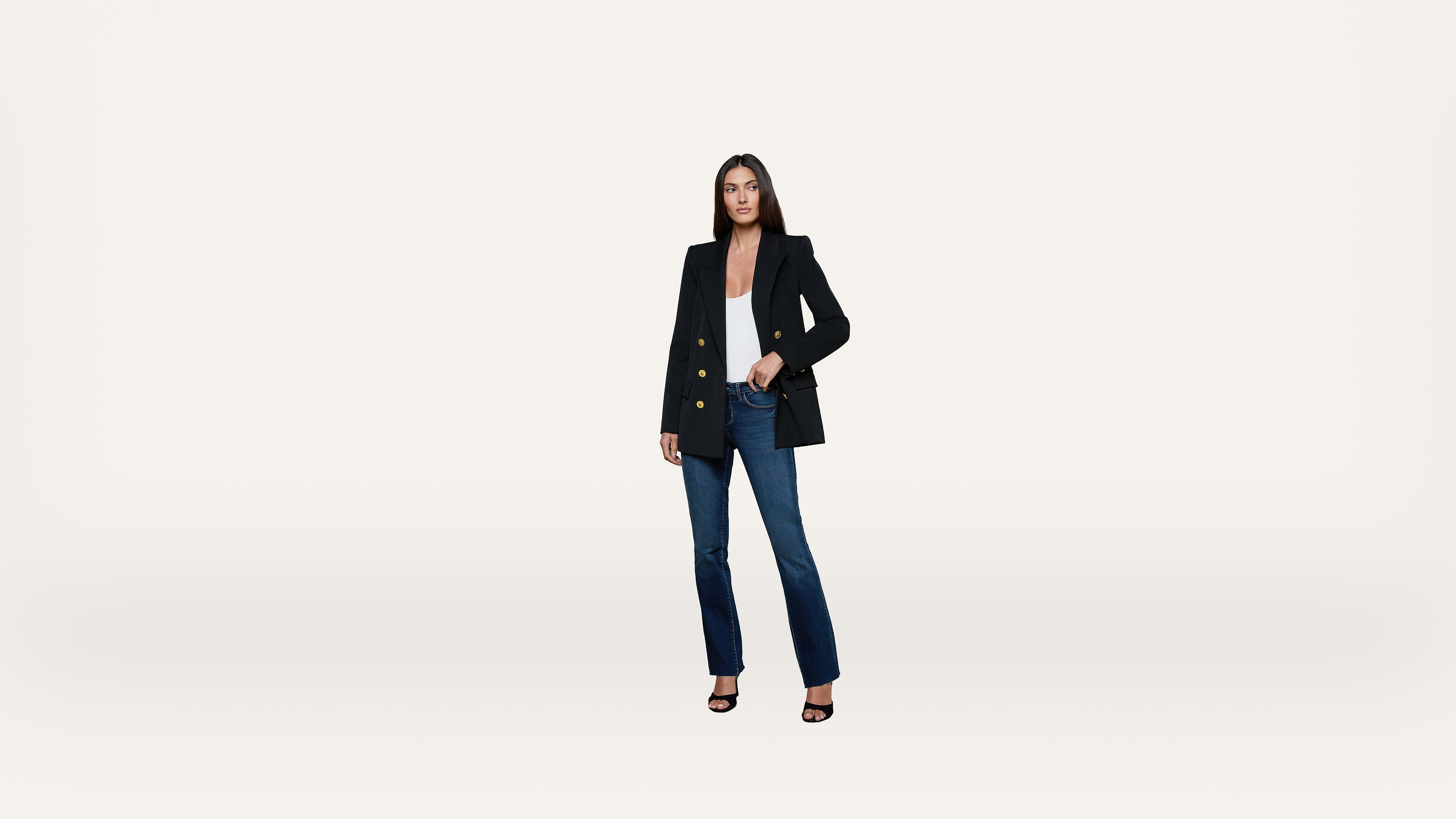 A woman stands against a plain white background wearing a black blazer, white top, blue jeans, and black heeled sandals, with one hand in her pocket.