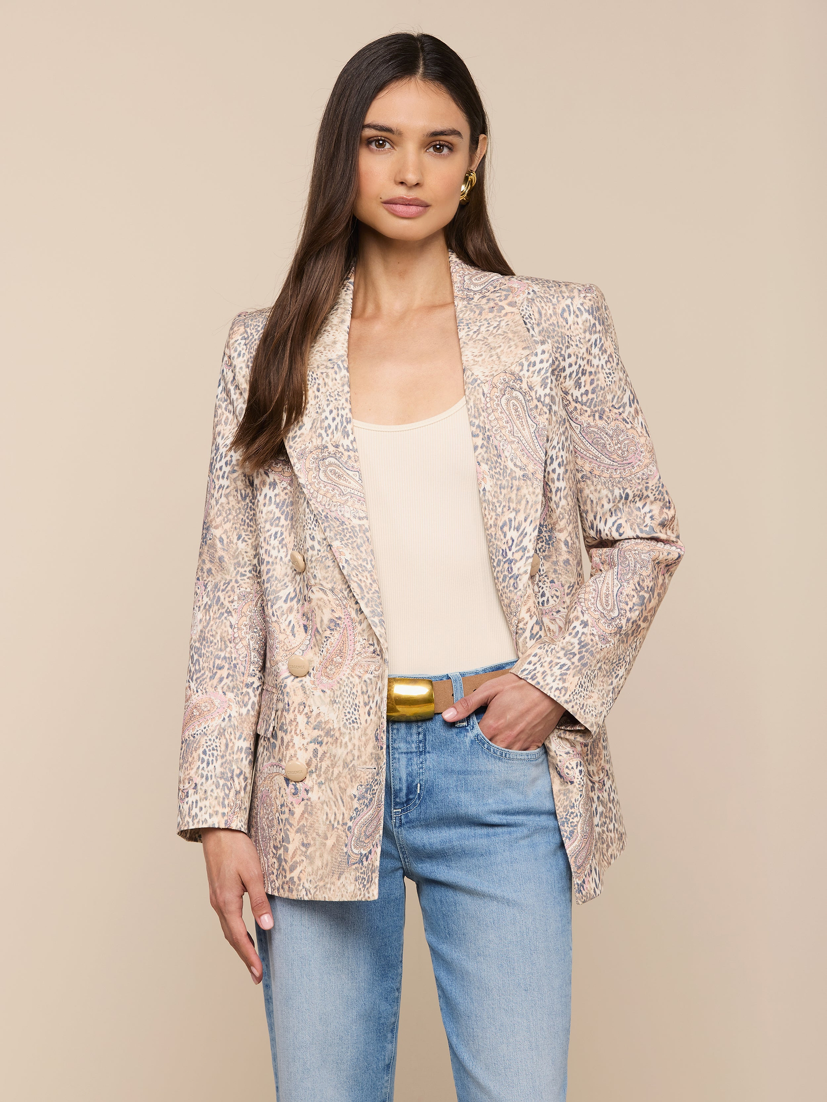 Una donna con lunghi capelli castani indossa il Cambrie Oversized Linen-Blend Blazer su un top beige, jeans blu chiaro e cintura dorata, su uno sfondo beige.