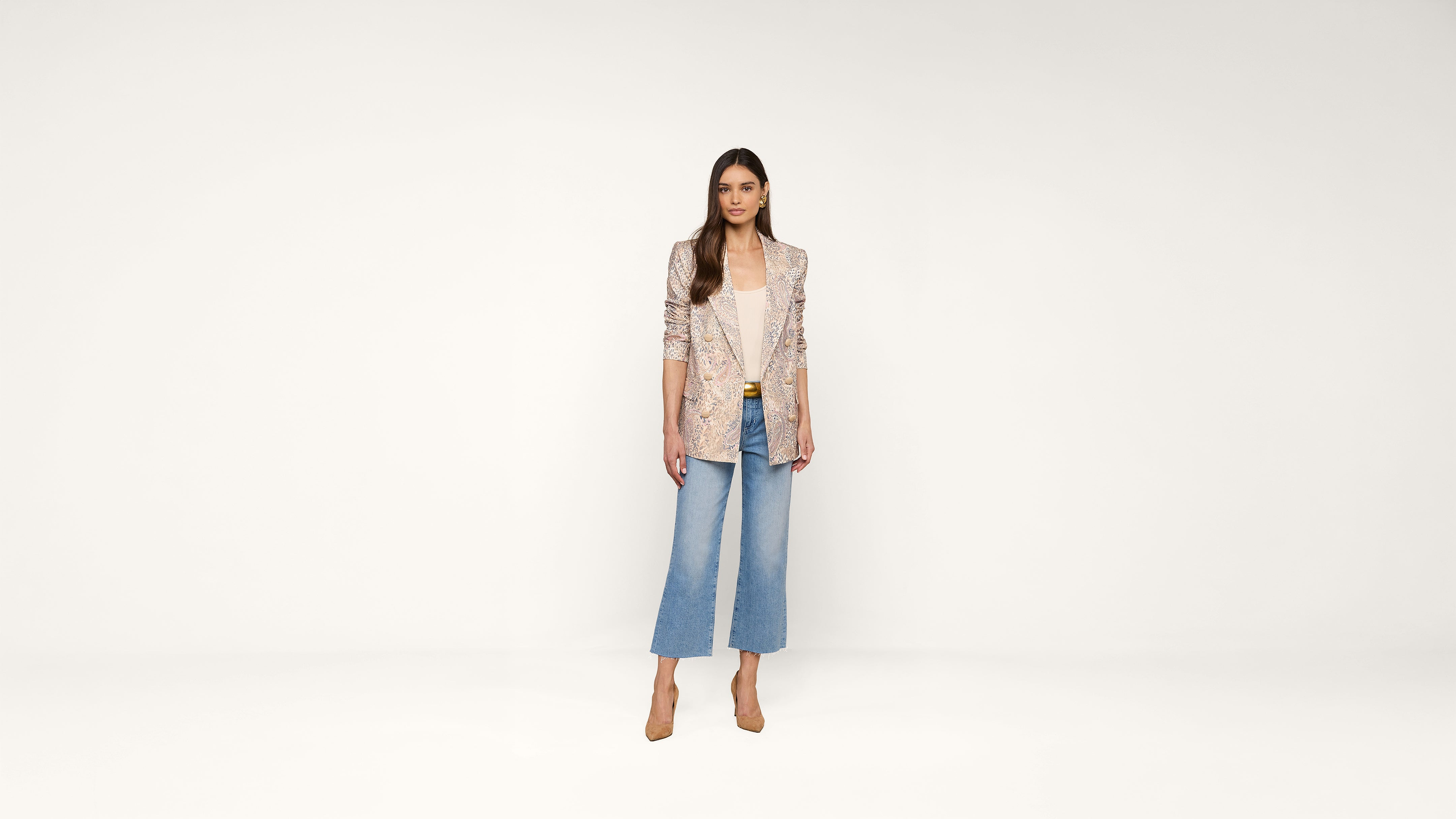 Woman standing against a plain white background, wearing a light patterned blazer, white top, wide-leg blue jeans, and tan heels. She has long brown hair and a neutral expression, with hands relaxed by her sides.