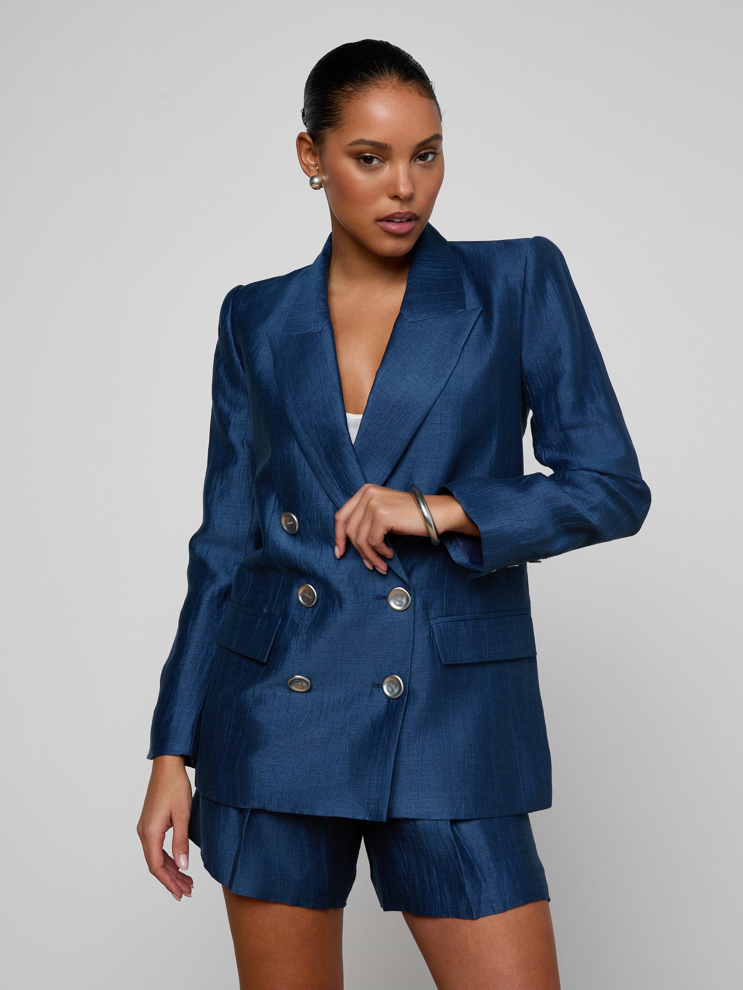 Wearing the Cambrie Oversized Blazer and matching shorts, a woman stands against a plain light background, gazing slightly to her left with one hand resting on her waist.