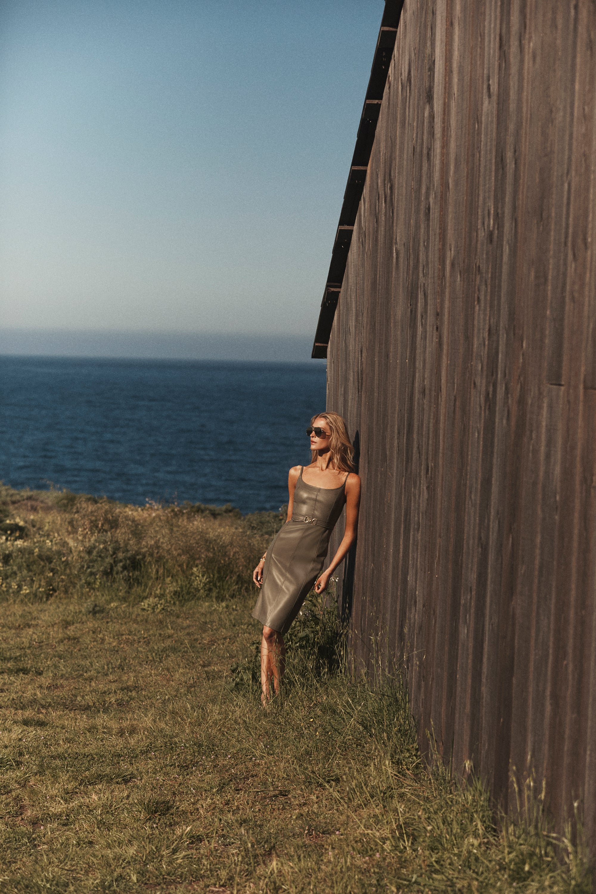 A woman in a light dress and sunglasses leans against a wooden wall, with a grassy area and the ocean visible in the background under a clear, sunny sky.