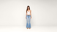 A woman wearing a white tank top, light blue flared jeans, and beige shoes stands facing forward against a plain white background.