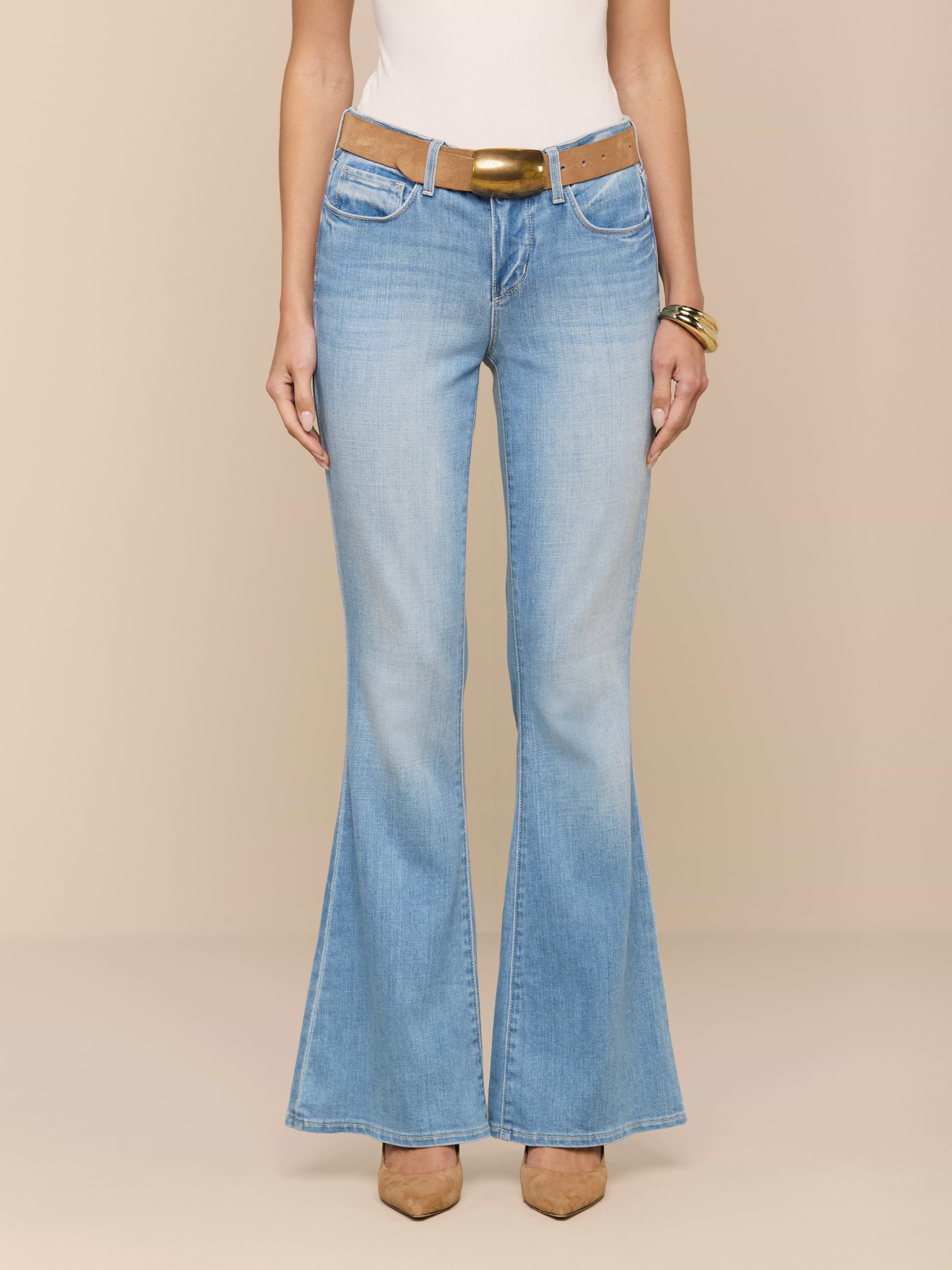 A person wearing the Taney Low-Rise Flare Jean in a light indigo wash, paired with a tan belt, white top, and brown pointed-toe shoes, stands against a plain white background with only their torso and legs visible.