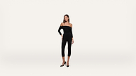 A woman wearing a black off-the-shoulder jumpsuit and black heels stands against a plain white background.