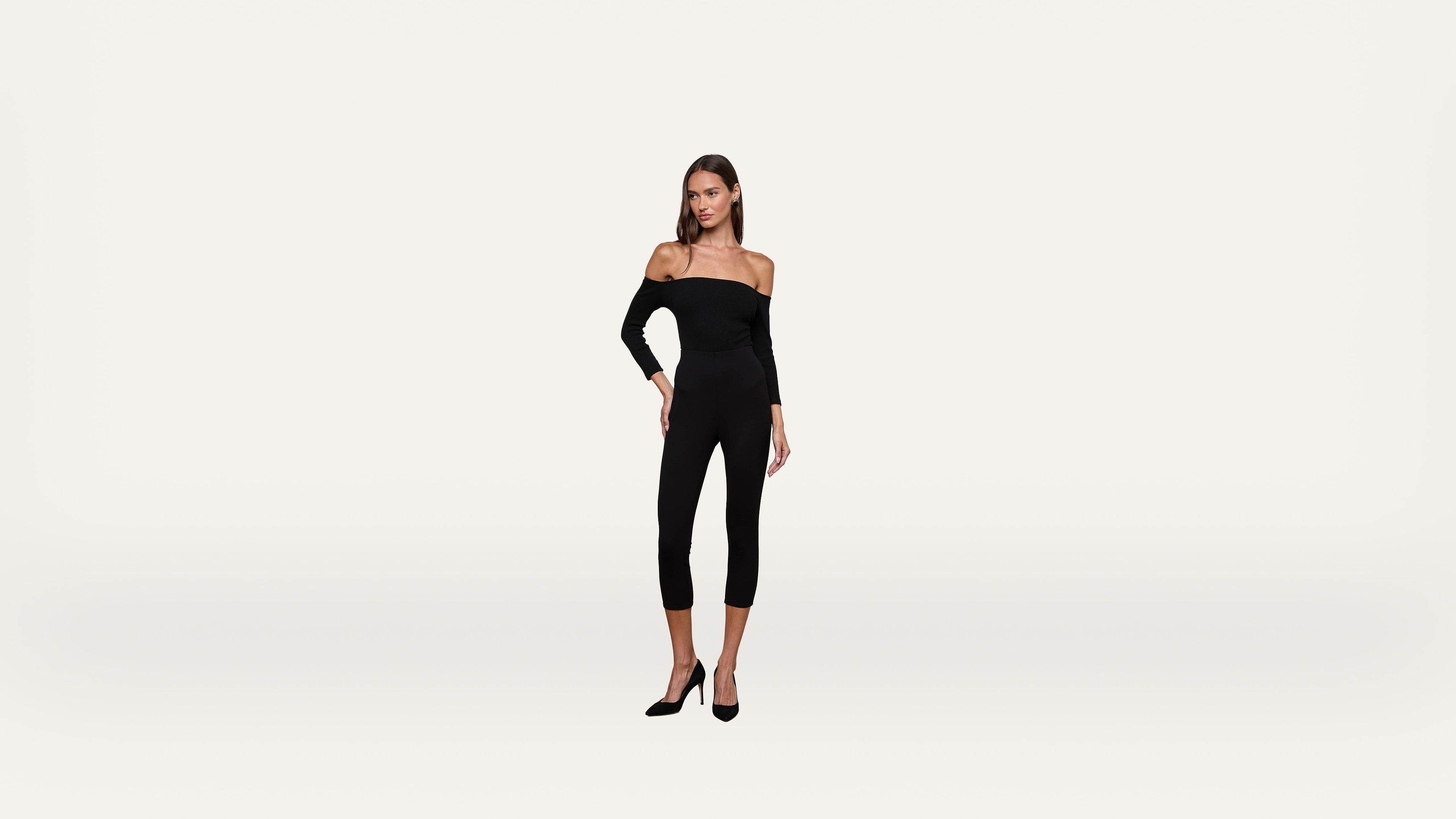 A woman wearing a black off-the-shoulder jumpsuit and black heels stands against a plain white background.