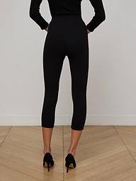 A person wearing the Dee Capri Pant in black, styled with a black long-sleeve top and black heels, stands facing away on a plain light background.