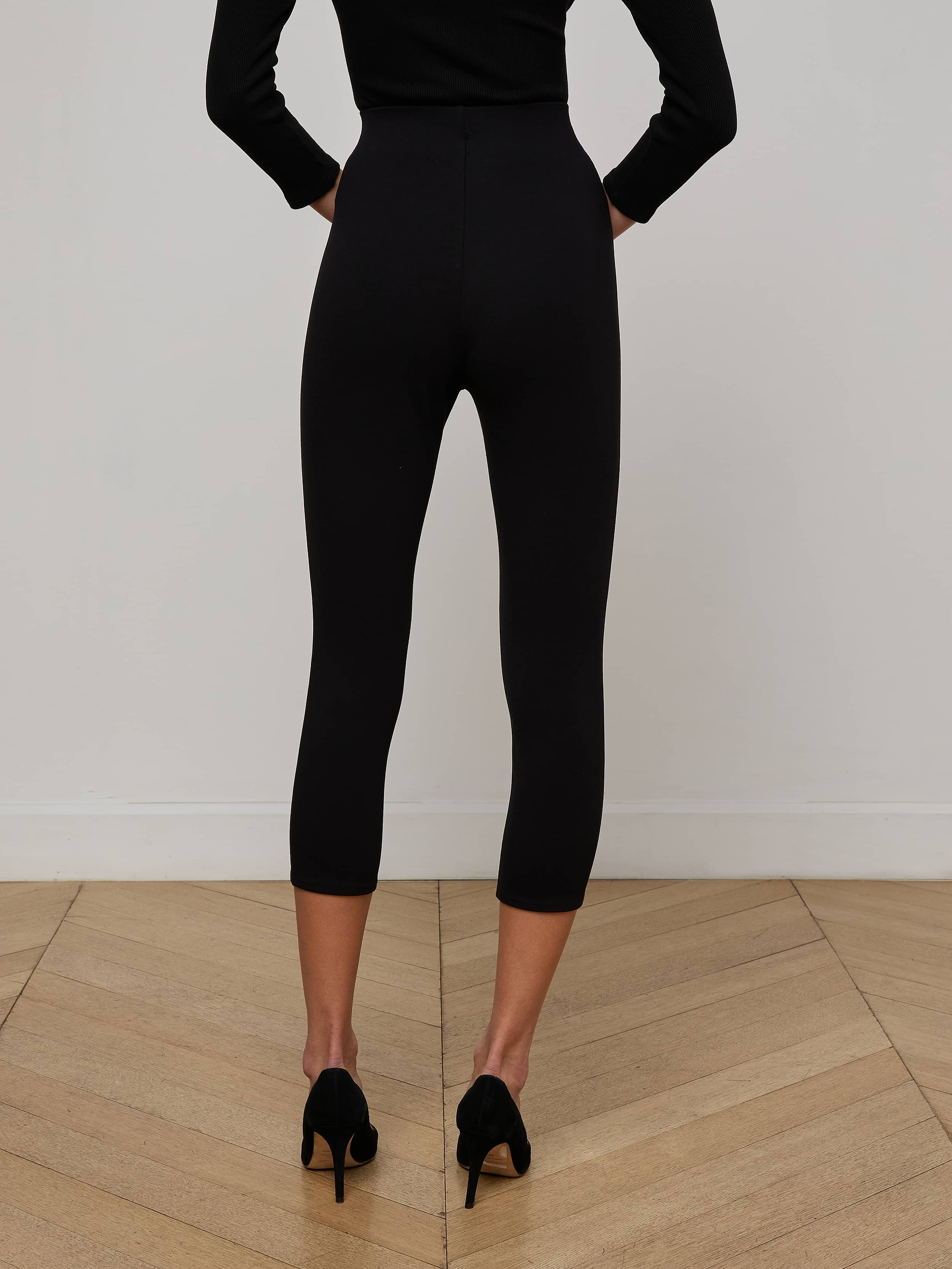 A person wearing the Dee Capri Pant in black, styled with a black long-sleeve top and black heels, stands facing away on a plain light background.