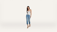 A woman with long dark hair is wearing a white tank top, blue jeans, and heels, walking confidently against a plain white background.