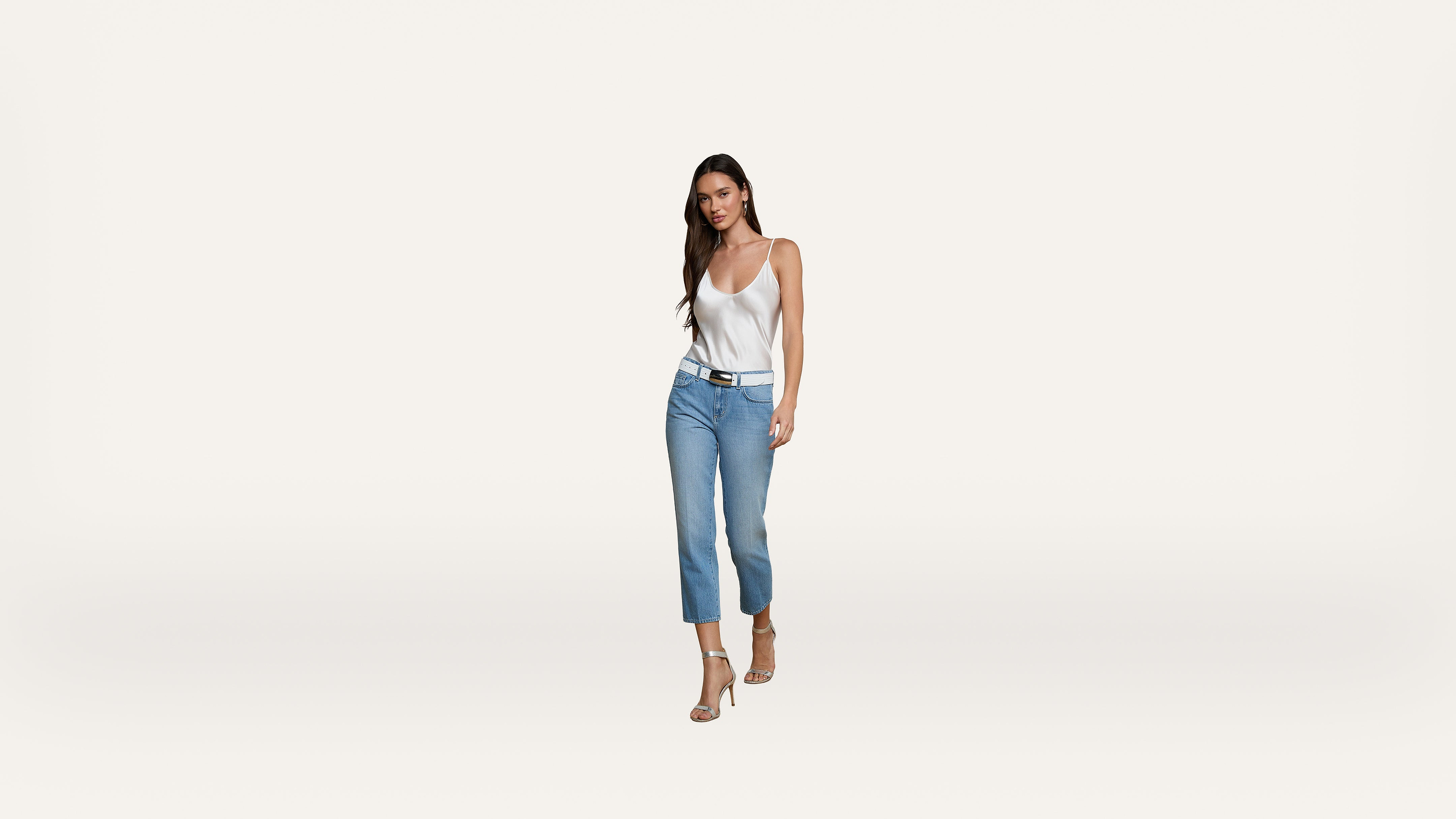 A woman with long dark hair is wearing a white tank top, blue jeans, and heels, walking confidently against a plain white background.