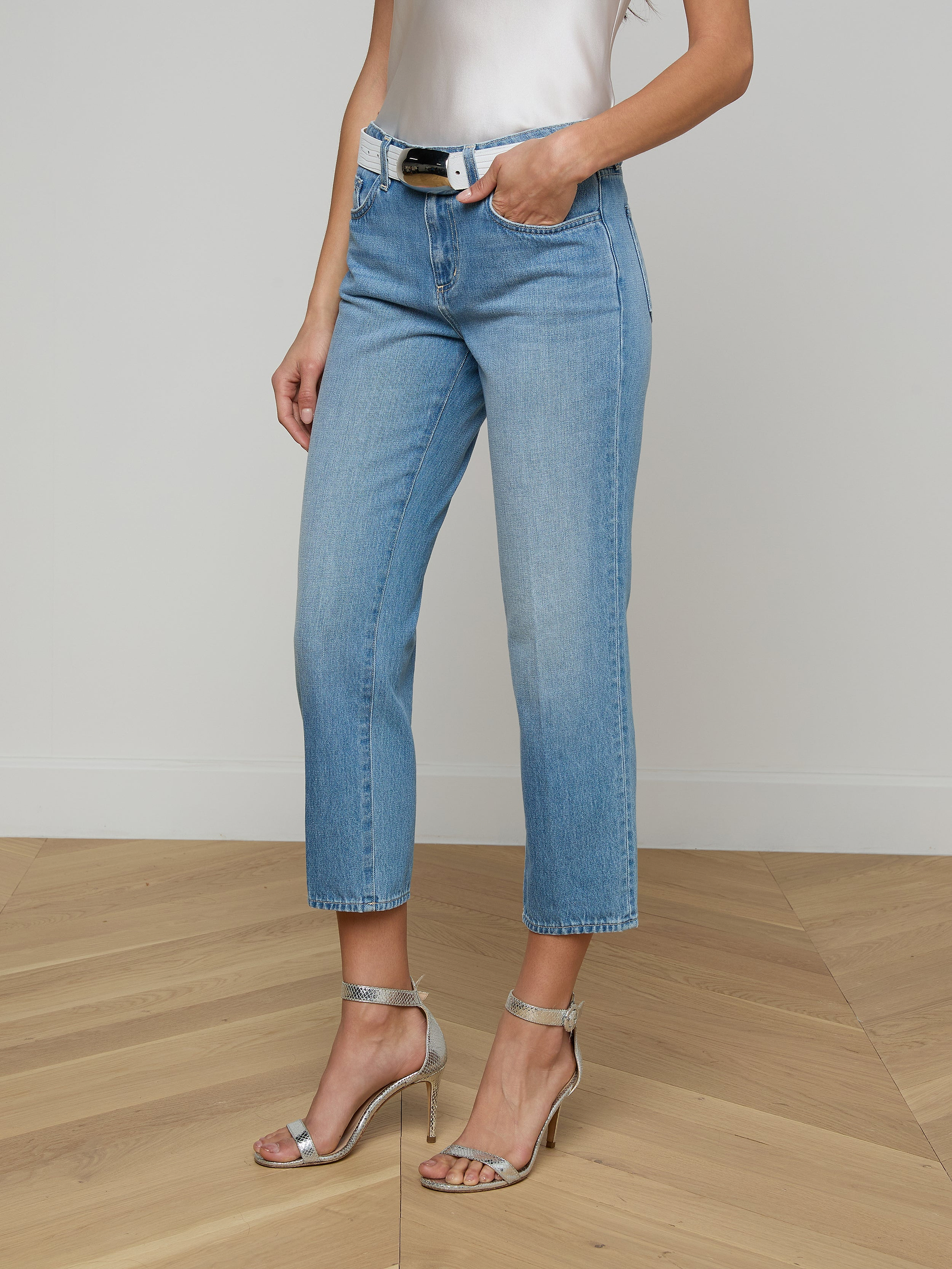 A woman stands with one hand in her pocket against a plain background, wearing the Thalina Cropped Stovepipe Jean in a light blue wash, paired with a white sleeveless top, black belt, and silver high-heeled sandals.