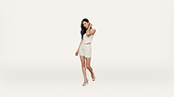 A woman wearing a light-colored, sleeveless top and matching skirt with heels stands against a plain white background, looking down and touching her hair.