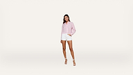 Woman in a light pink button-up shirt and white shorts poses against a plain white background, wearing white heeled sandals and standing with one hand in her pocket.