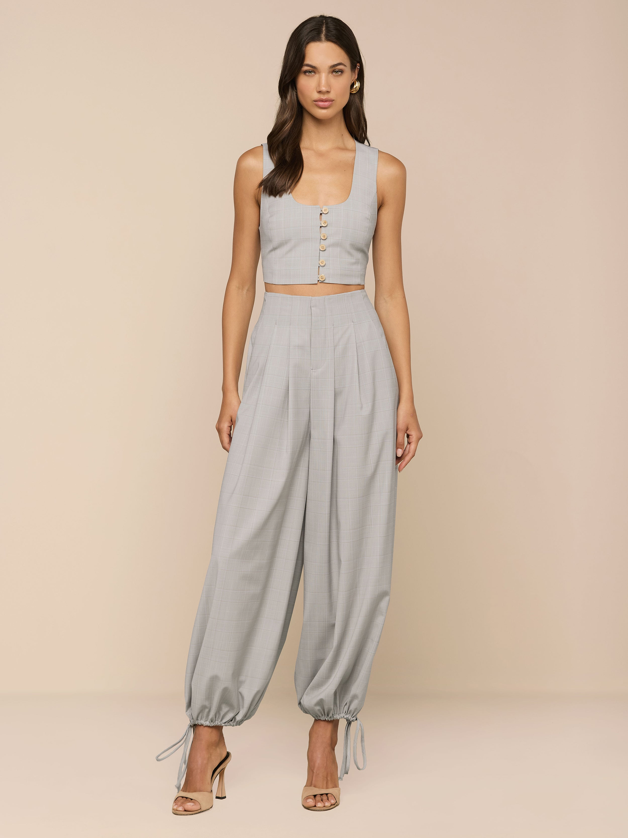 A woman stands against a plain background, wearing a light gray sleeveless crop top with buttons and matching Ysola Pant—ultra-high rise, wide-leg pants with adjustable hems. She completes the look with open-toe heels.