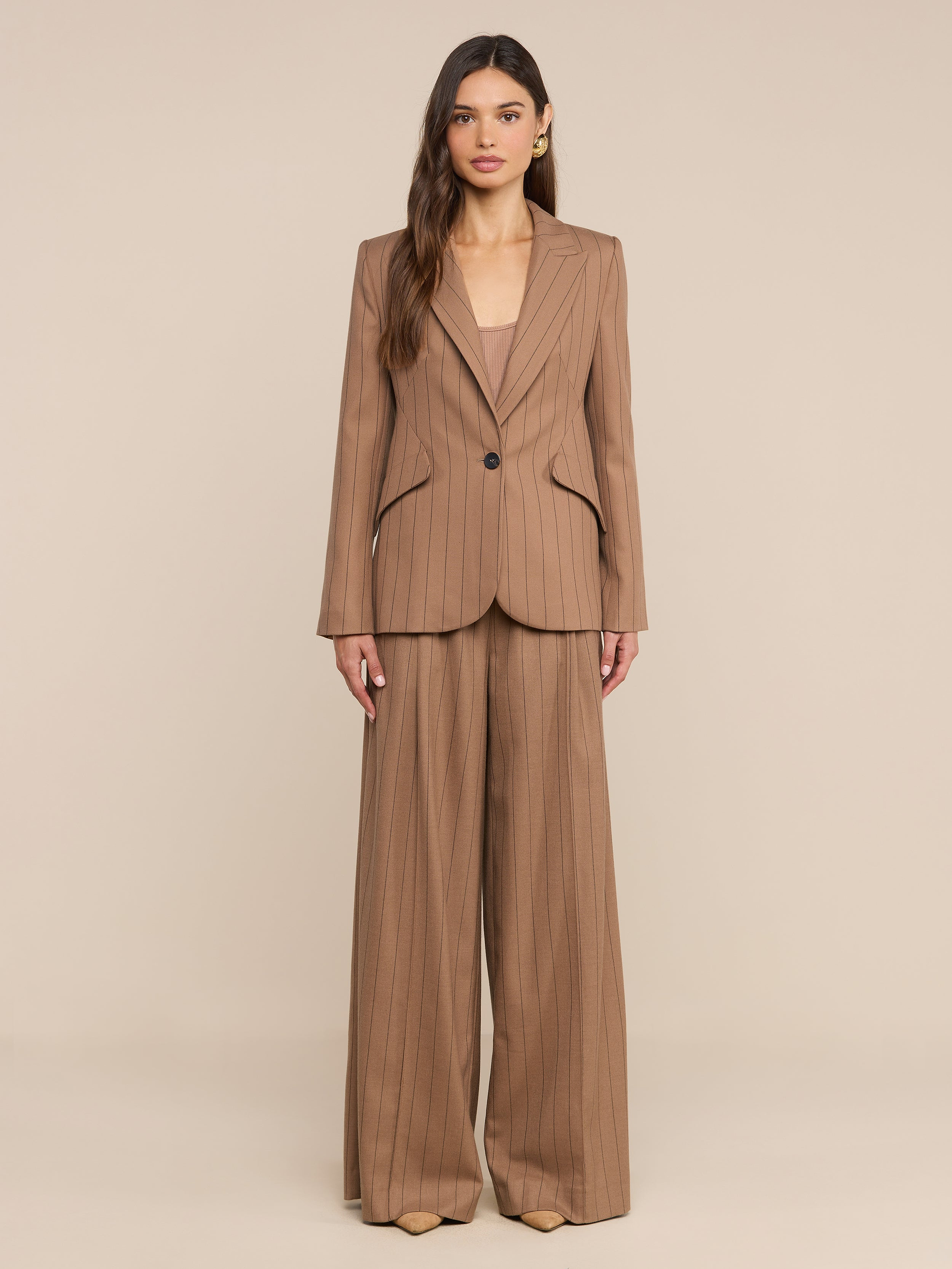 A woman stands facing forward against a plain white background, wearing a tan pinstripe suit featuring the Connie Drawstring Trouser—a high-rise, wide-leg style paired with a matching blazer.