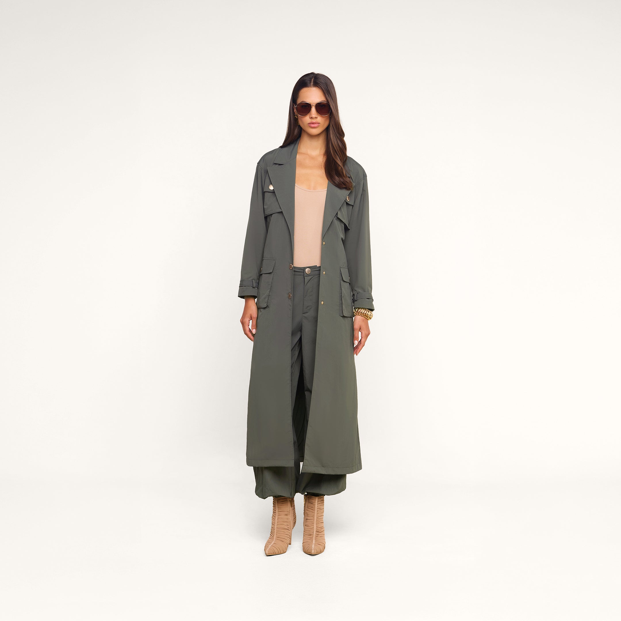 A woman wearing sunglasses, a long ash olive green trench coat with cargo pockets, the Greer Nylon Jogger in olive, a beige top, and tan heeled boots stands against a plain white background.