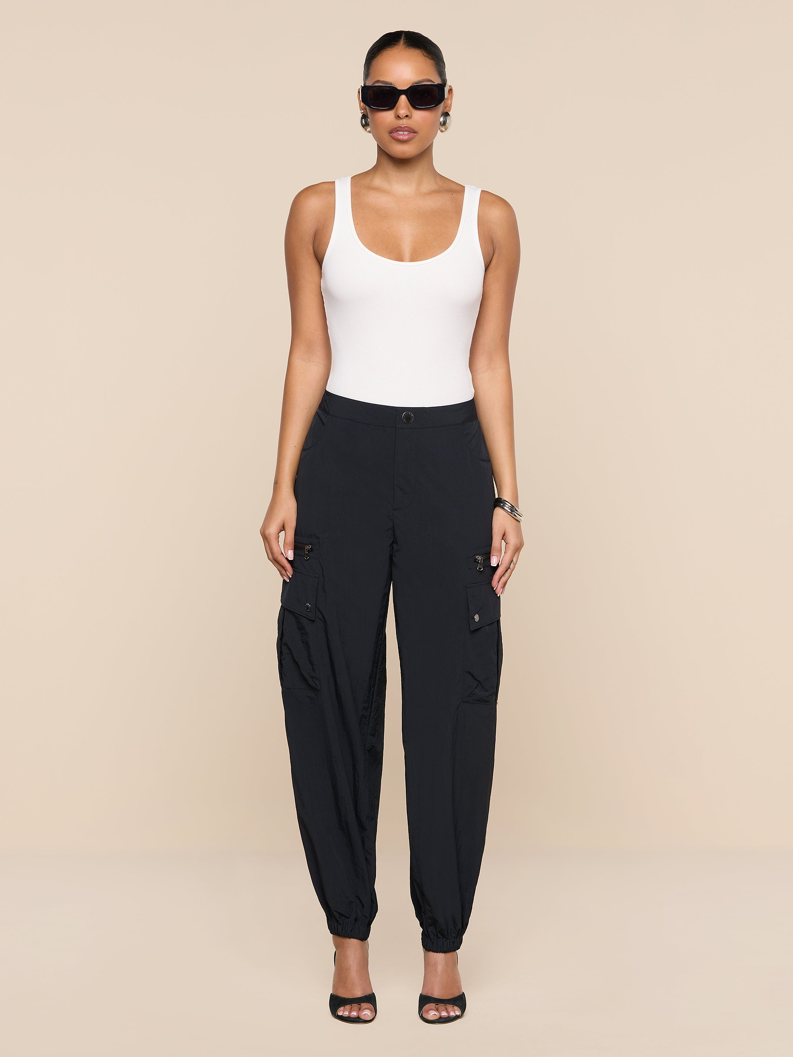 A woman wearing sunglasses, a white tank top, the Greer Nylon Jogger with cargo pockets in black, and black heeled sandals stands against a plain white background.