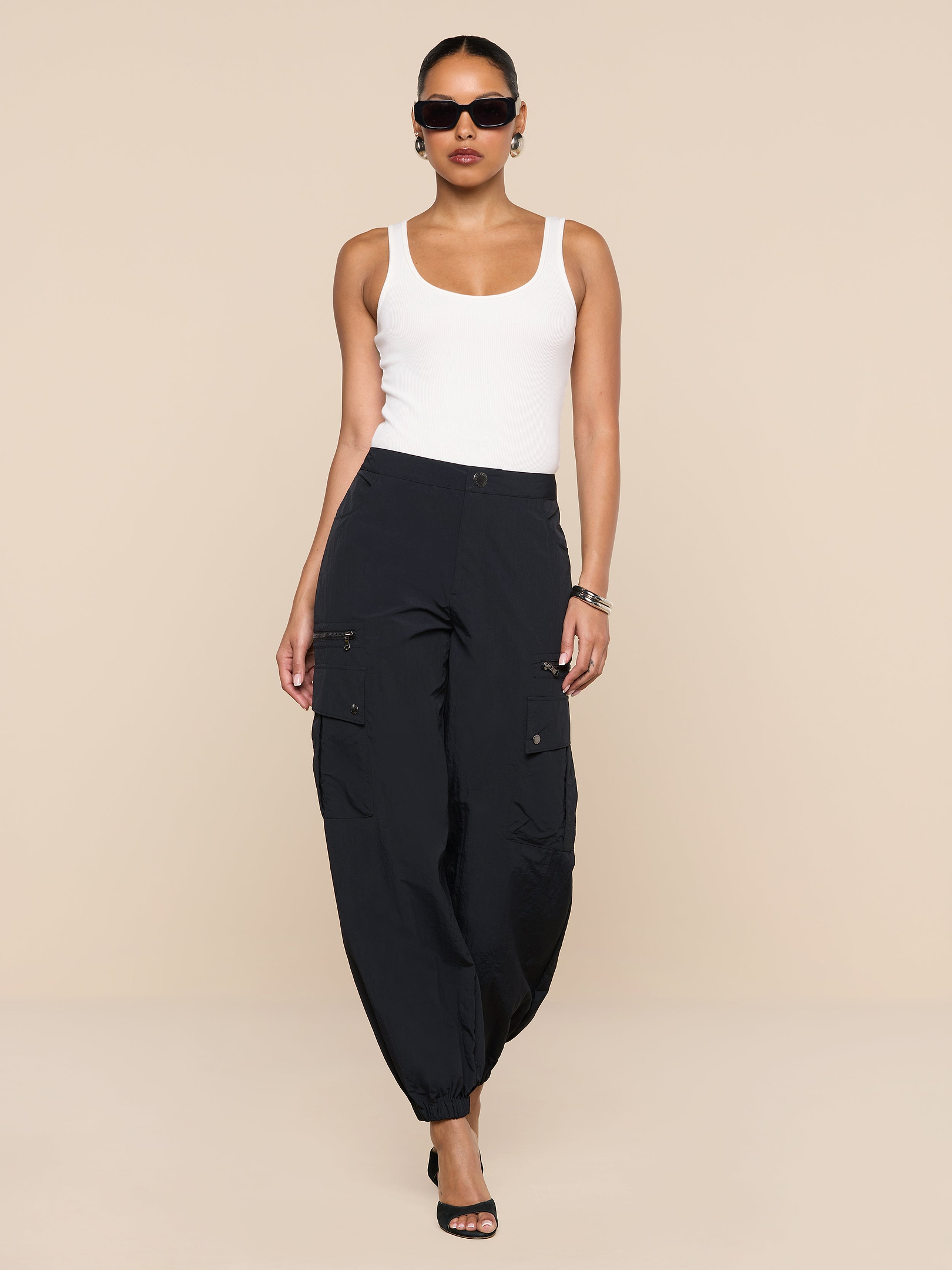 A woman stands confidently against a plain white background, wearing a white tank top, the Greer Nylon Jogger with cargo pockets in black, black heels, and sunglasses.