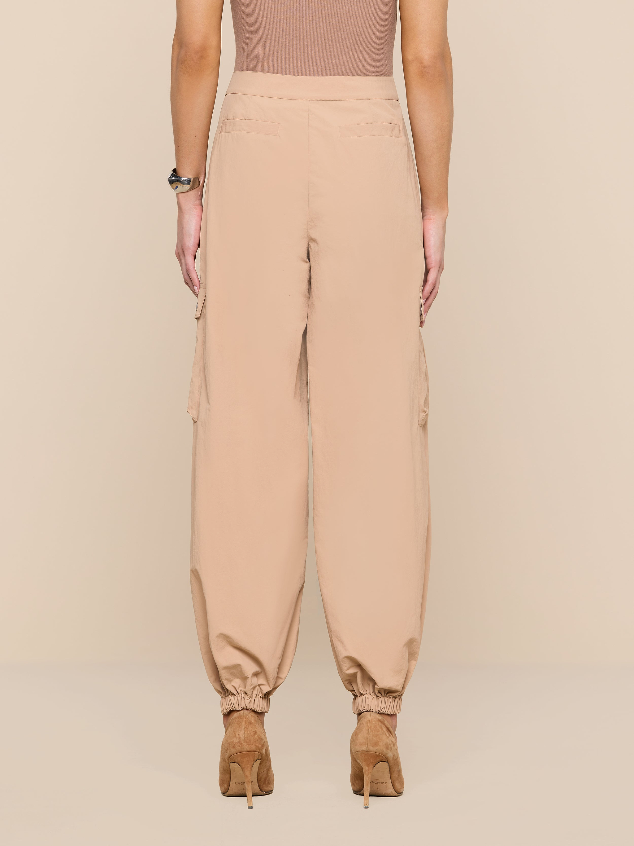 A person wearing the Greer Nylon Jogger—high-waisted, loose beige pants with cinched ankles—paired with tan heeled shoes, shown from the back against a plain white background.