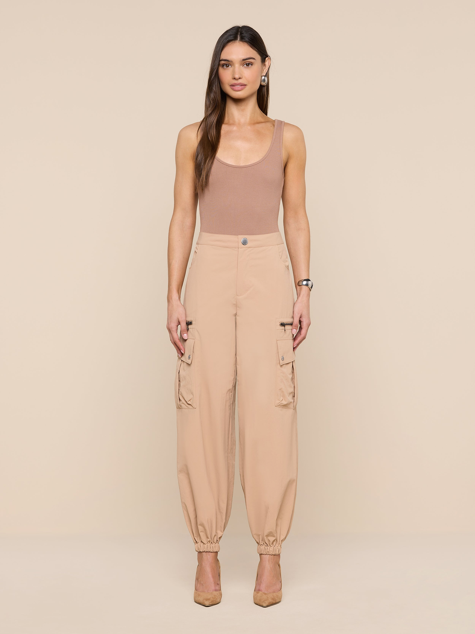 A woman stands facing forward against a plain white background, wearing a tan sleeveless top, the Greer Nylon Jogger—high-waisted beige cargo pants with cargo pockets—and beige heels.