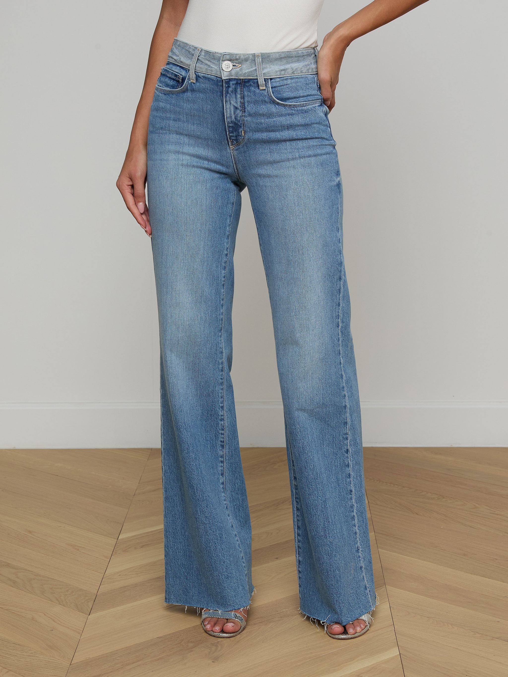 A woman models the Louane Wide-Leg Flare Jean in light blue, featuring a high-rise fit and frayed hems, paired with a white sleeveless top. She stands against a plain light background with her hands at her sides and her face out of view.