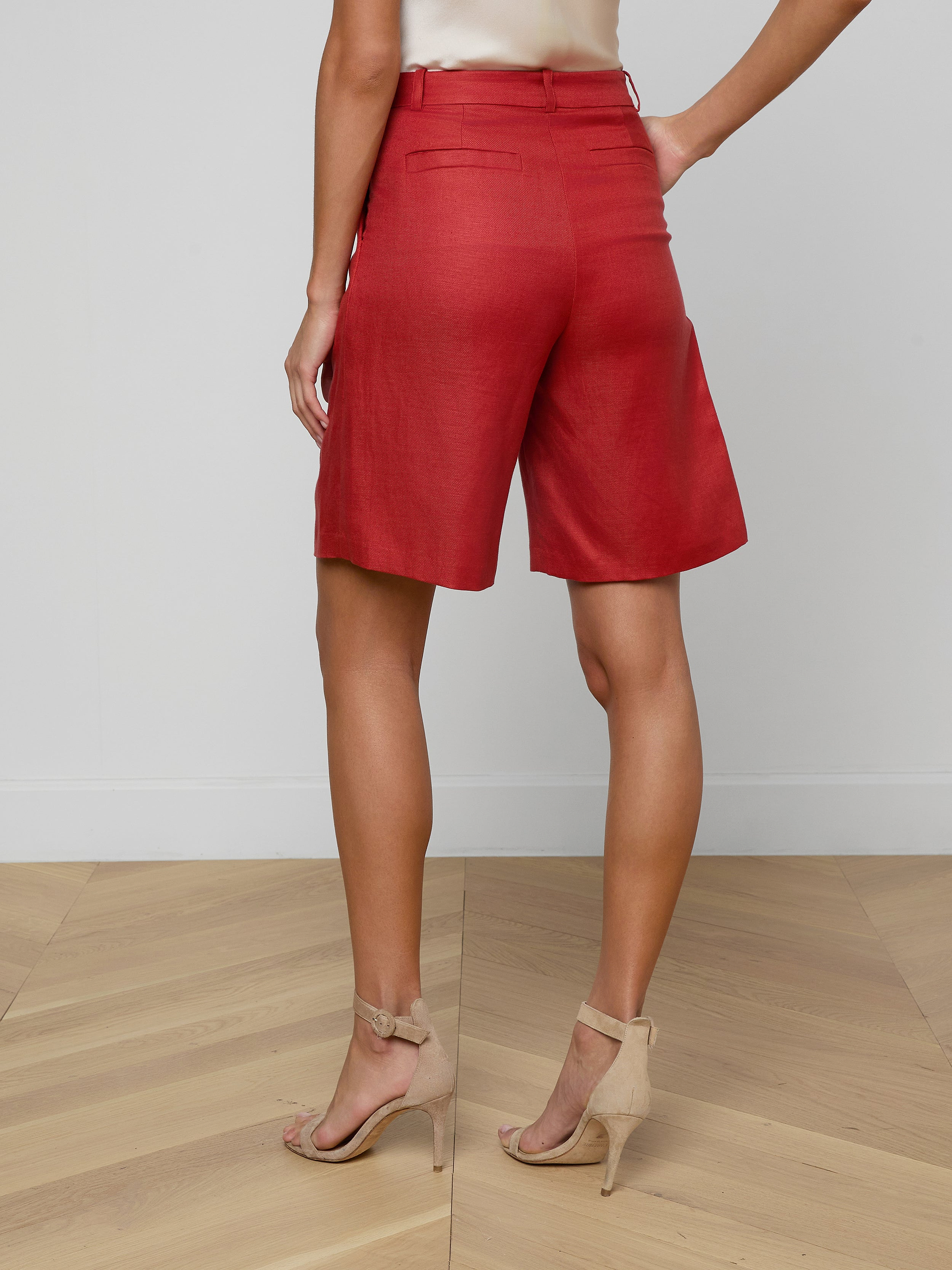 A woman stands with her back to the camera, wearing the Doretta Bermuda Short in a red linen blend, paired with a light-colored top and beige high-heeled sandals against a plain white background—perfect for summer-in-the-office style.