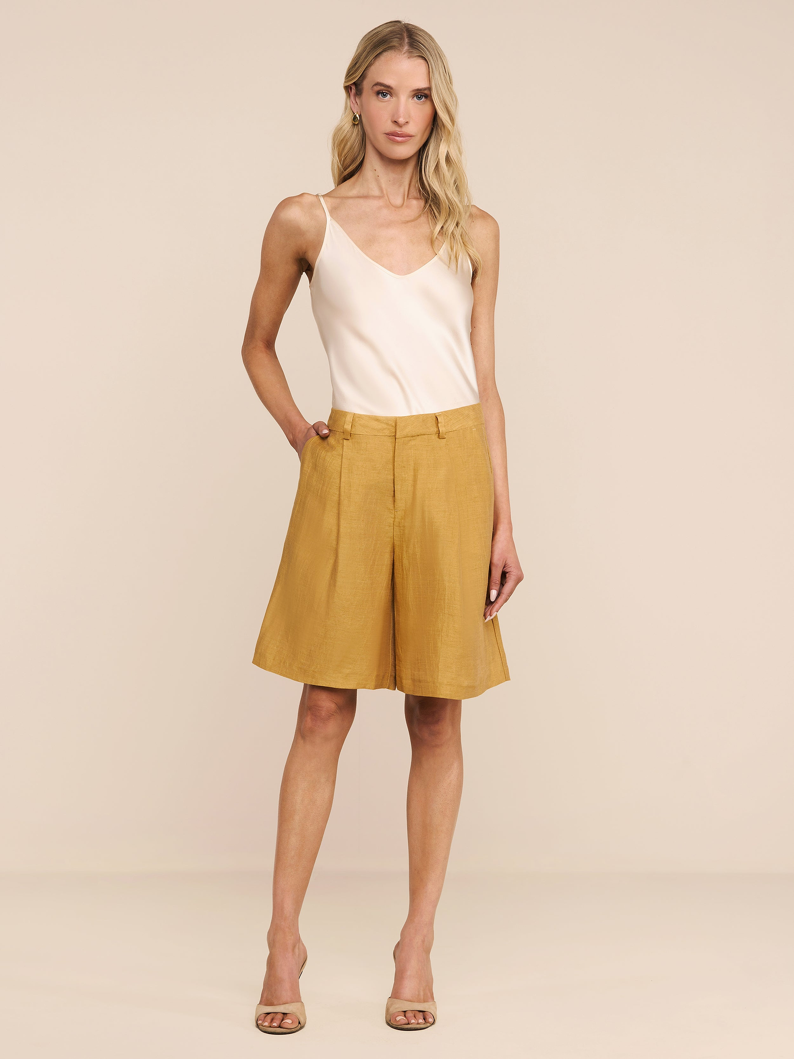 Una donna in canotta color crema, tacchi beige e bermuda Doretta Linen-Blend si staglia su uno sfondo chiaro con una mano in tasca.