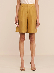 A person stands against a white background wearing the Doretta Linen-Blend Bermuda Short in gold, paired with a light beige top and open-toed heels. Their arms are relaxed by their sides.