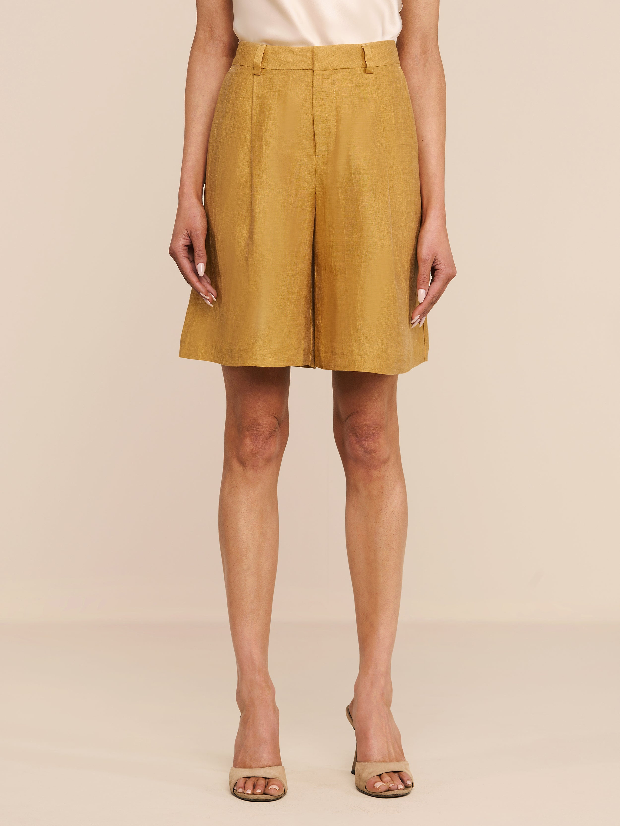 A person stands against a white background wearing the Doretta Linen-Blend Bermuda Short in gold, paired with a light beige top and open-toed heels. Their arms are relaxed by their sides.