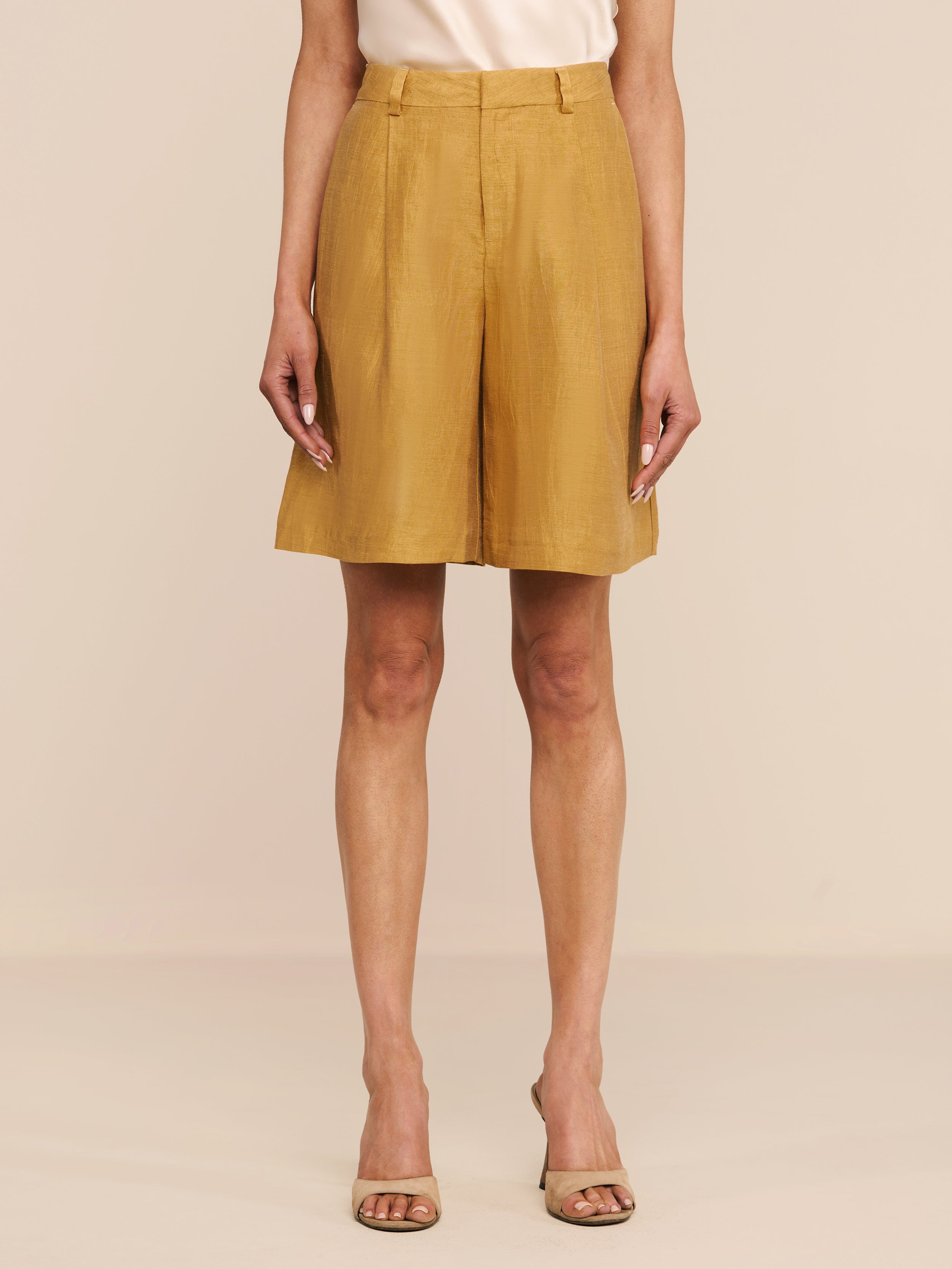 A person stands against a white background wearing the Doretta Linen-Blend Bermuda Short in gold, paired with a light beige top and open-toed heels. Their arms are relaxed by their sides.