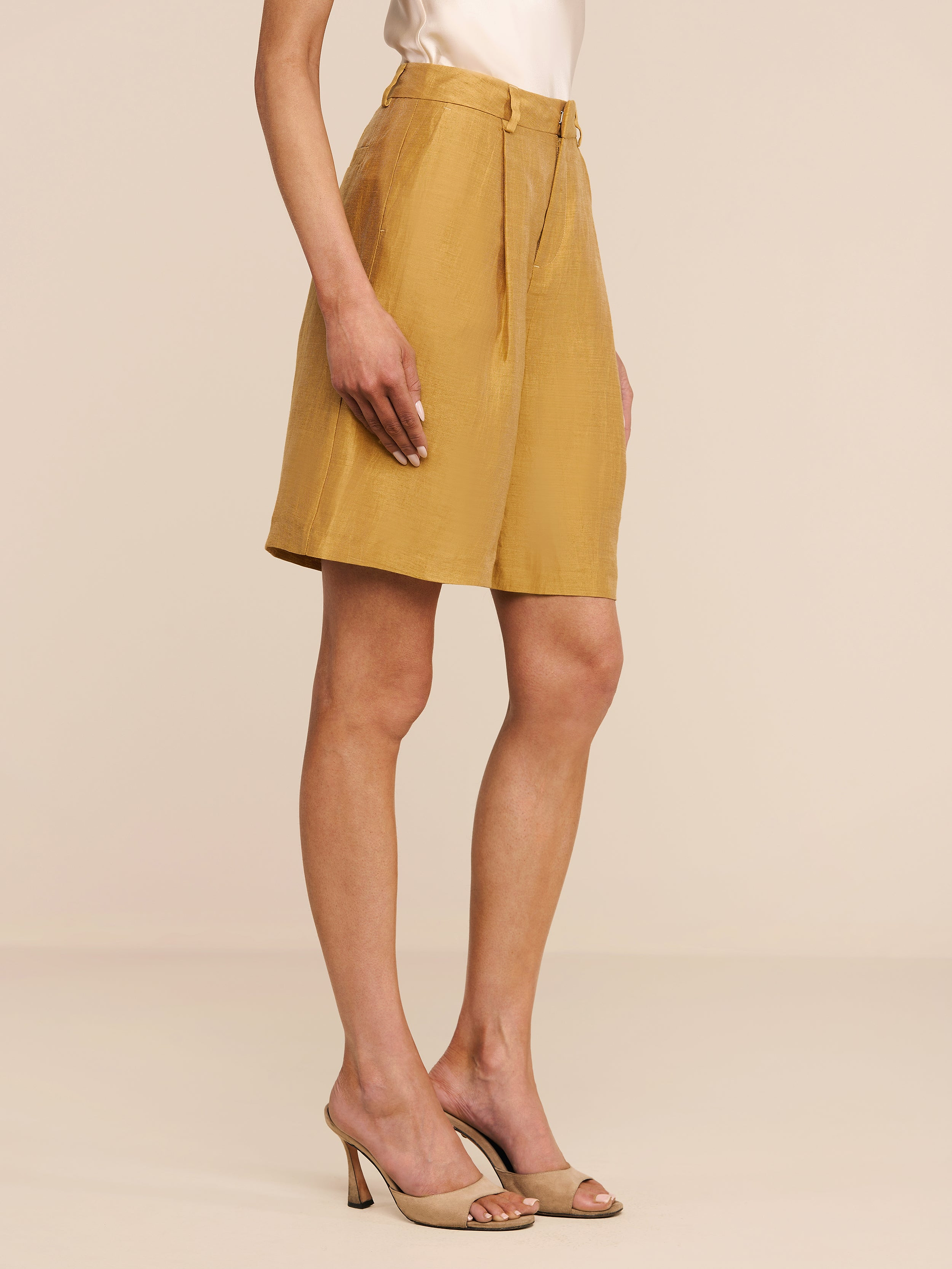 A woman stands on a white background wearing the Doretta Linen-Blend Bermuda Short in gold, paired with a light beige sleeveless top and beige high-heeled sandals. Her arms are relaxed by her side.
