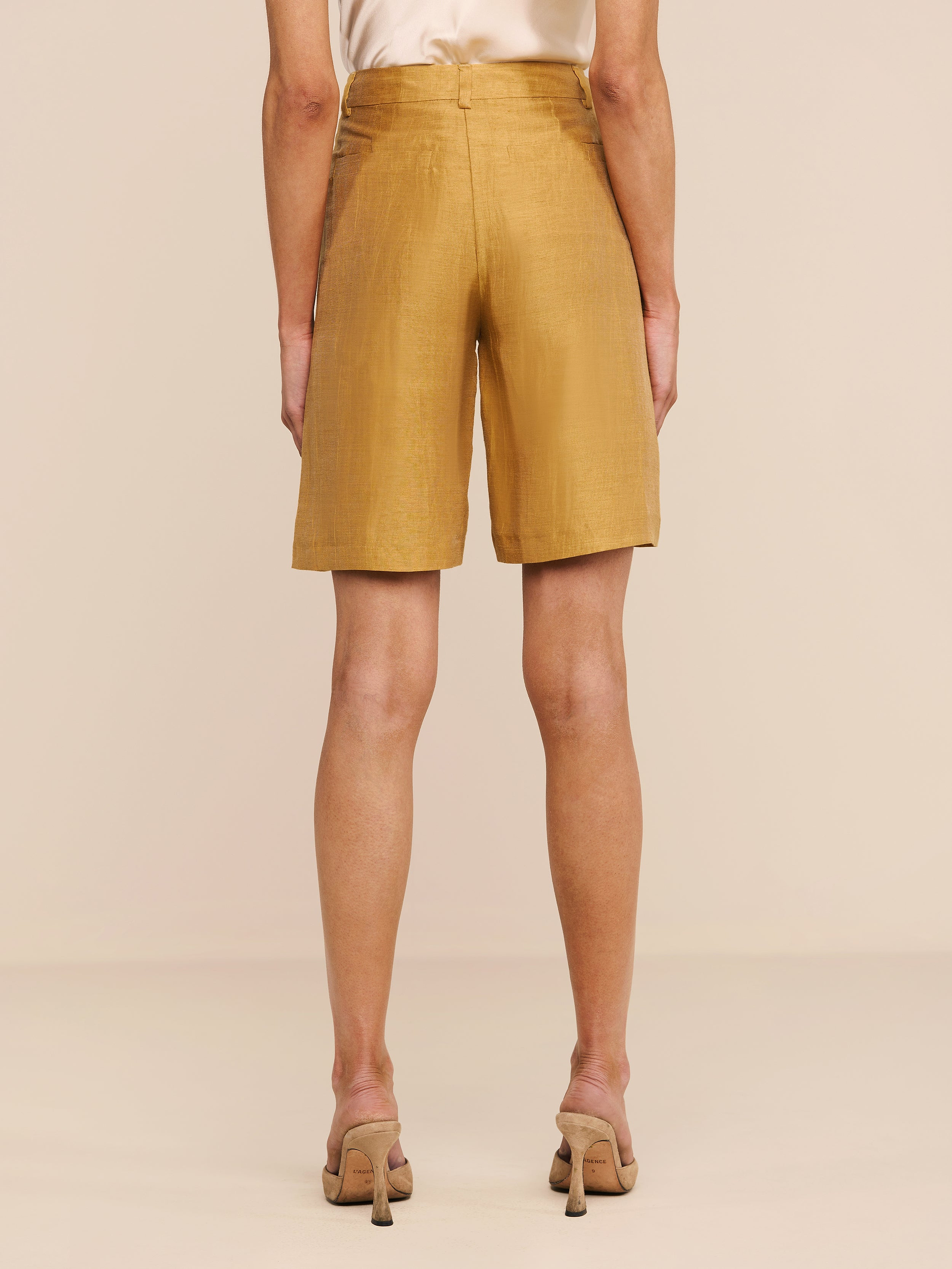 Wearing the Doretta Linen-Blend Bermuda Short in mustard yellow and beige heeled sandals, a person is shown from the back standing against a plain white background.
