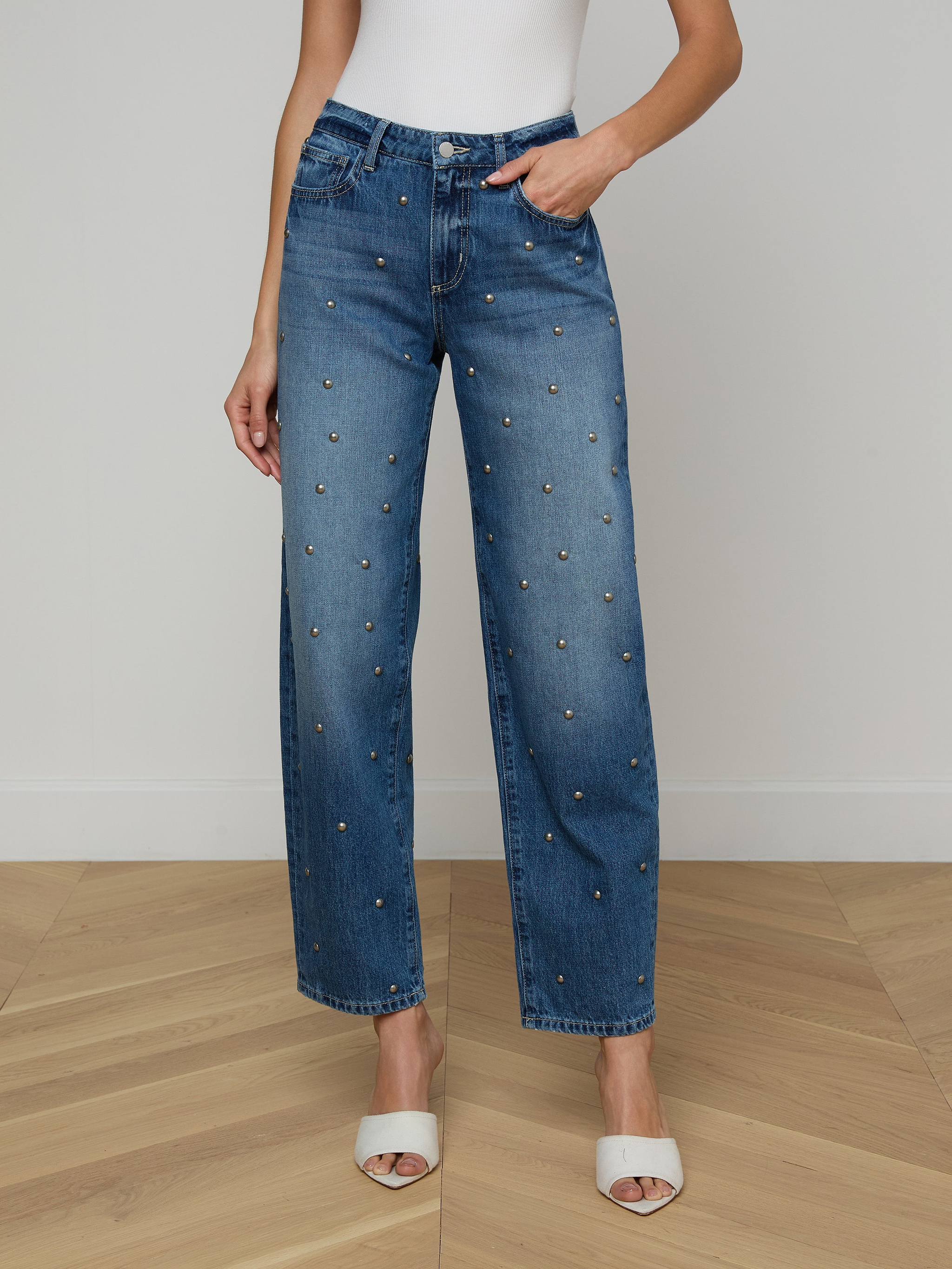 A woman wears the Bex Studded Rodeo Jean—high-rise, wide-leg denim featuring small studs—paired with a white sleeveless top and white open-toe heels, standing against a plain background.