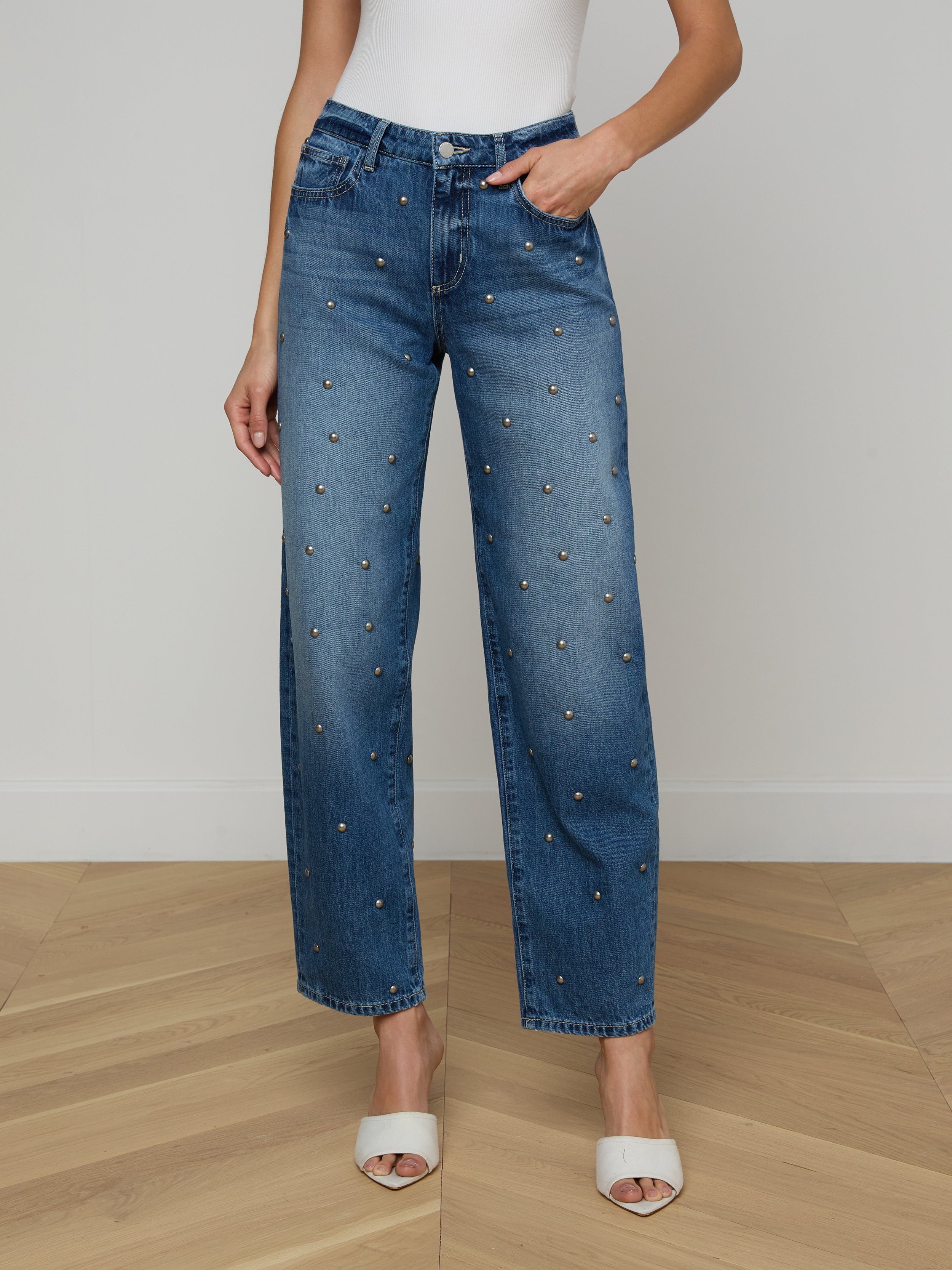 A woman wears the Bex Studded Rodeo Jean—high-rise, wide-leg denim featuring small studs—paired with a white sleeveless top and white open-toe heels, standing against a plain background.
