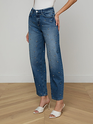 A person is wearing the Bex Studded Rodeo Jean, a pair of high rise blue jeans with small embellishments, styled with a white top and white open-toe heeled sandals, posed against a plain light background.