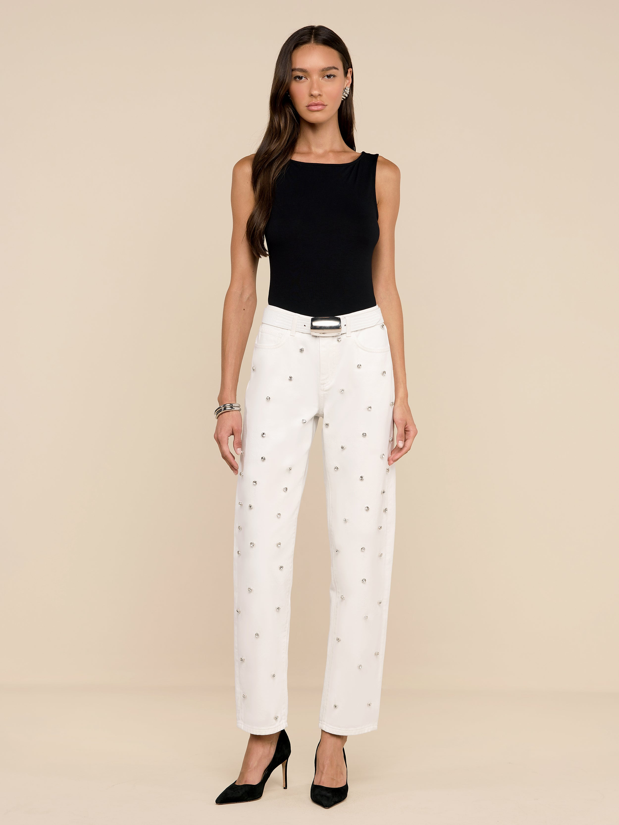 Une femme aux longs cheveux bruns se tient sur un fond beige et porte le Bex Embellished Rodeo Jean, un haut noir sans manches, une ceinture noire, des talons noirs et un bracelet. Elle a une expression neutre.