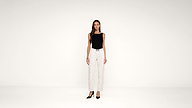 Woman standing in a minimalist studio, wearing a black sleeveless top, white high-waisted pants with a pattern, a black belt, and black heels against a plain white background.