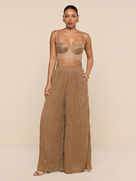 A woman stands against a plain white background wearing a beige crop top and the Ethel Pleated Pant, loose, high-waisted crinkle chiffon pants in brown, paired with sandals.