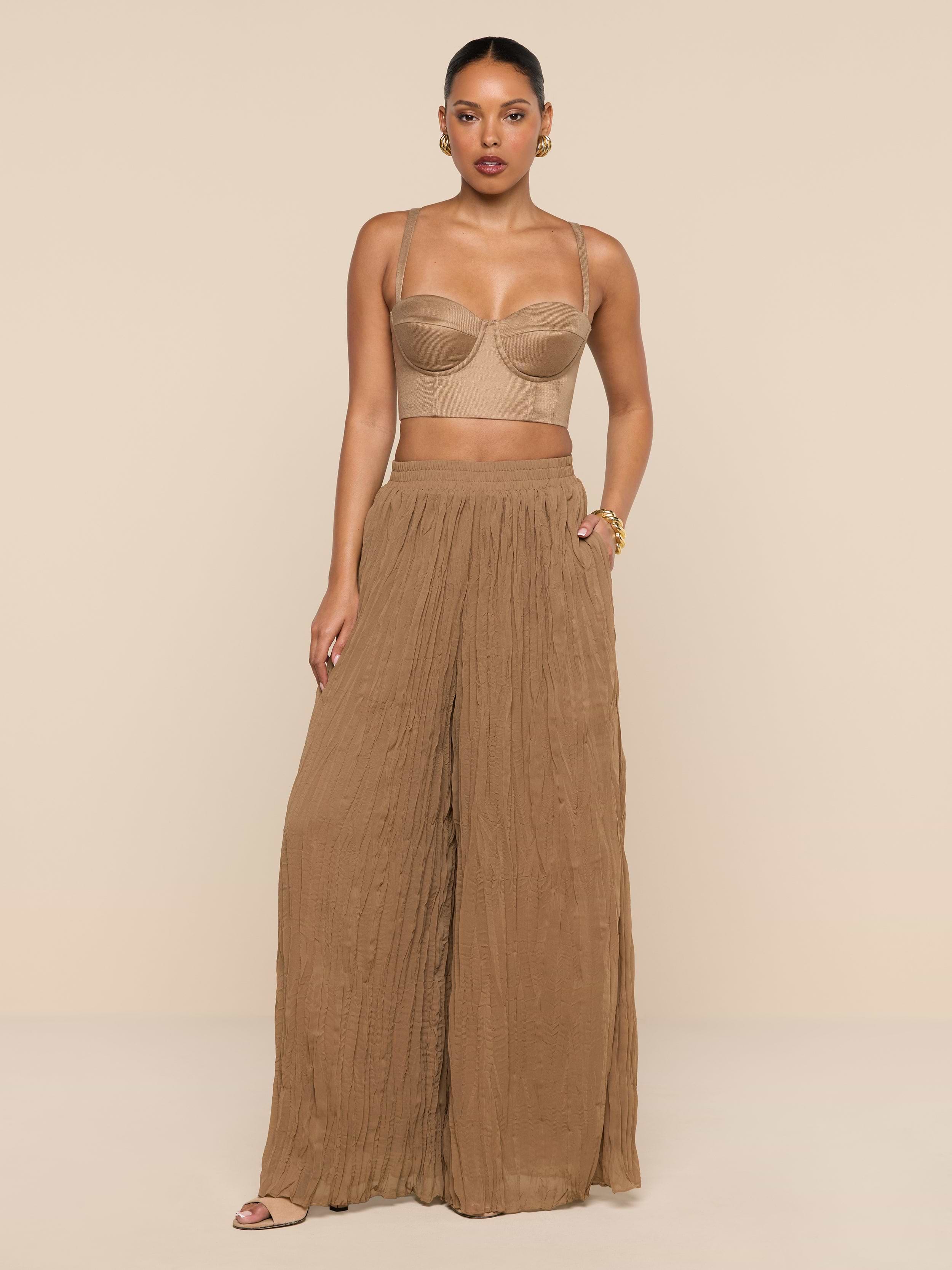 A woman stands against a plain white background wearing a beige crop top and the Ethel Pleated Pant, loose, high-waisted crinkle chiffon pants in brown, paired with sandals.
