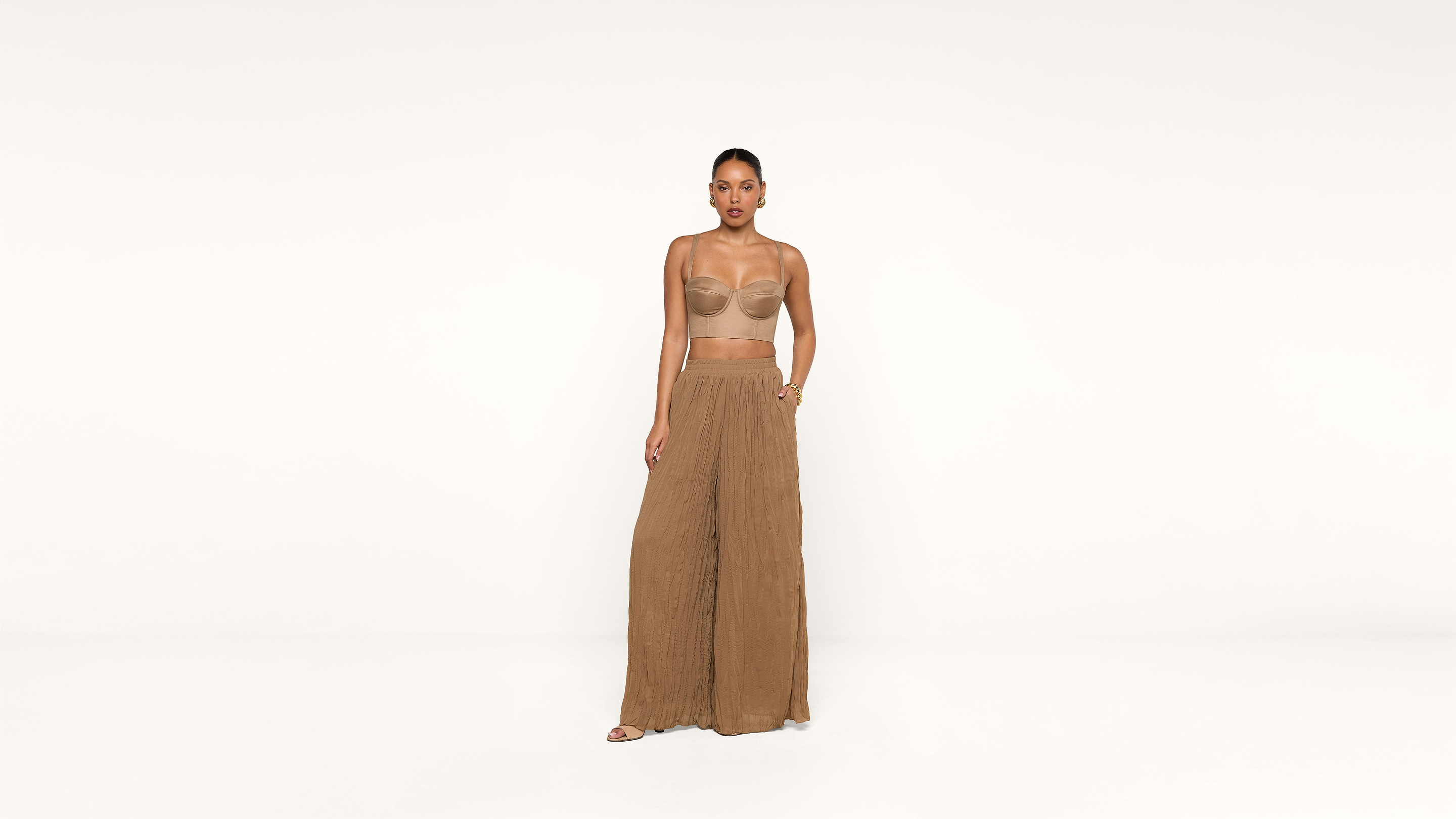A woman stands against a white background wearing a strapless nude bandeau top and loose-fitting, high-waisted brown pants, with her hair pulled back and one hand resting on her hip.