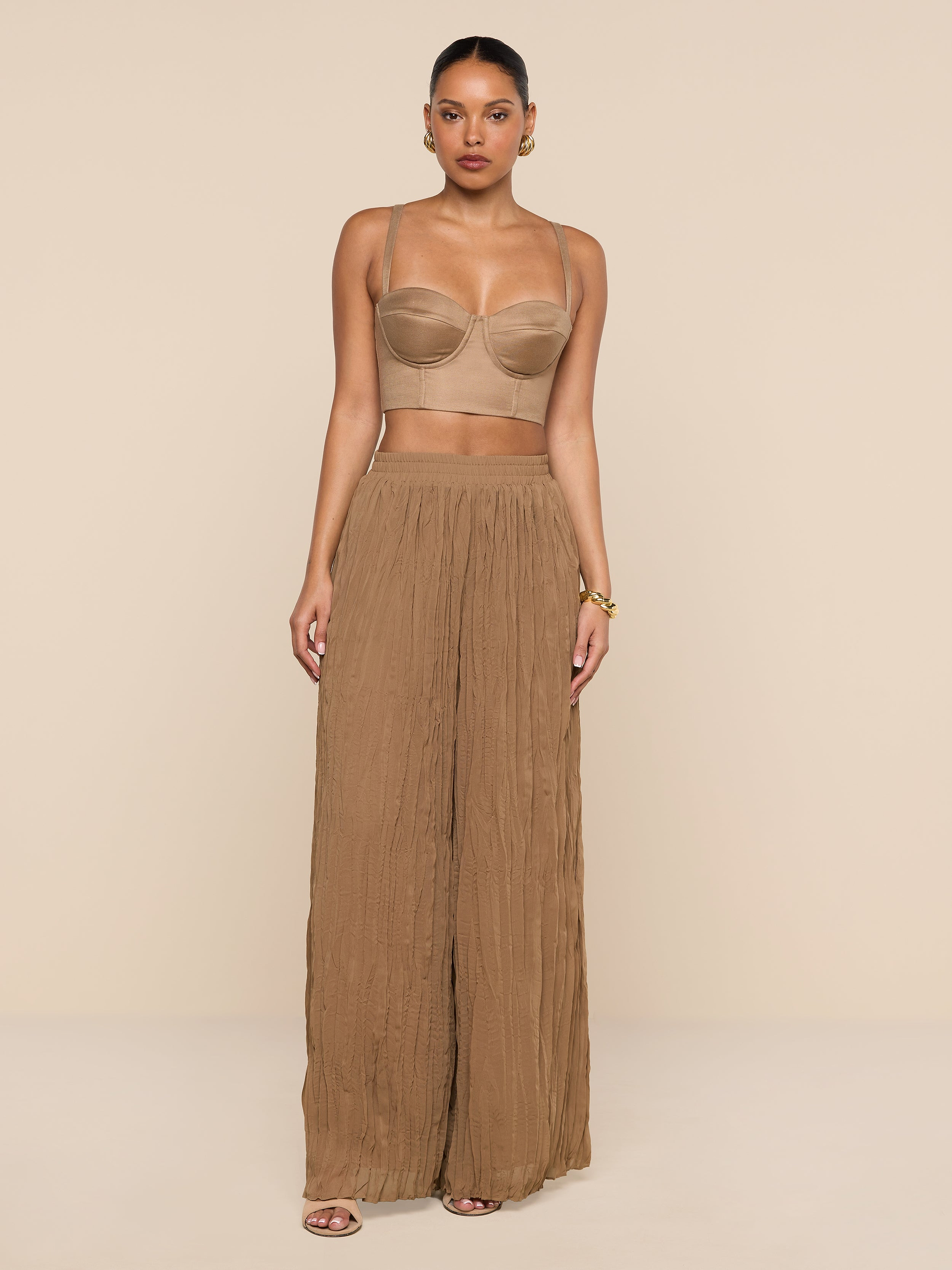 A woman stands against a plain white background wearing a tan bandeau top, the Ethel Pleated Pant—a sheer, wide-legged brown summer style—sandals, and gold hoop earrings. Her hair is pulled back and her hands rest relaxed at her sides.
